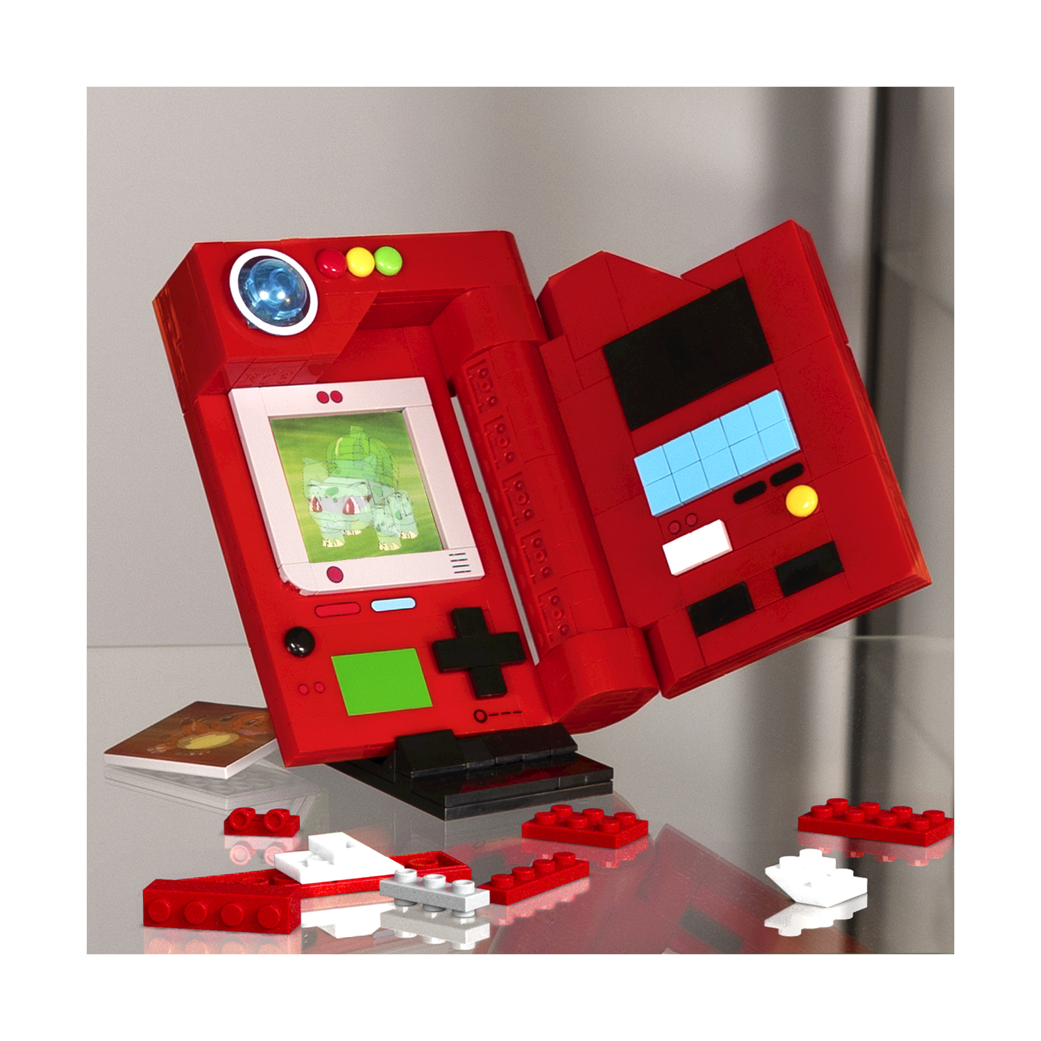 Czerwona konstrukcja Lego Game Boy, otwarta, z ekranem pokazującym Pokemona. Klocki Lego rozsiane.