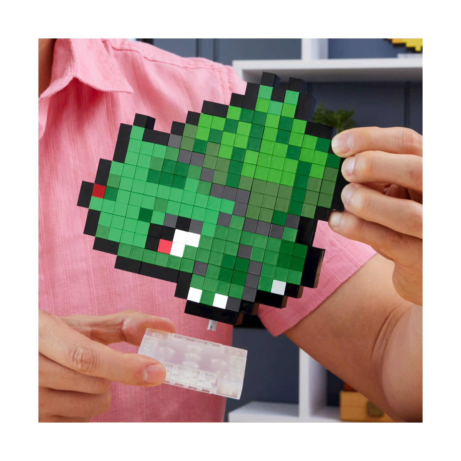 Una persona tiene in mano un drago verde pixelato. Il drago è fatto di blocchi, con contorni neri e una base di plastica trasparente.