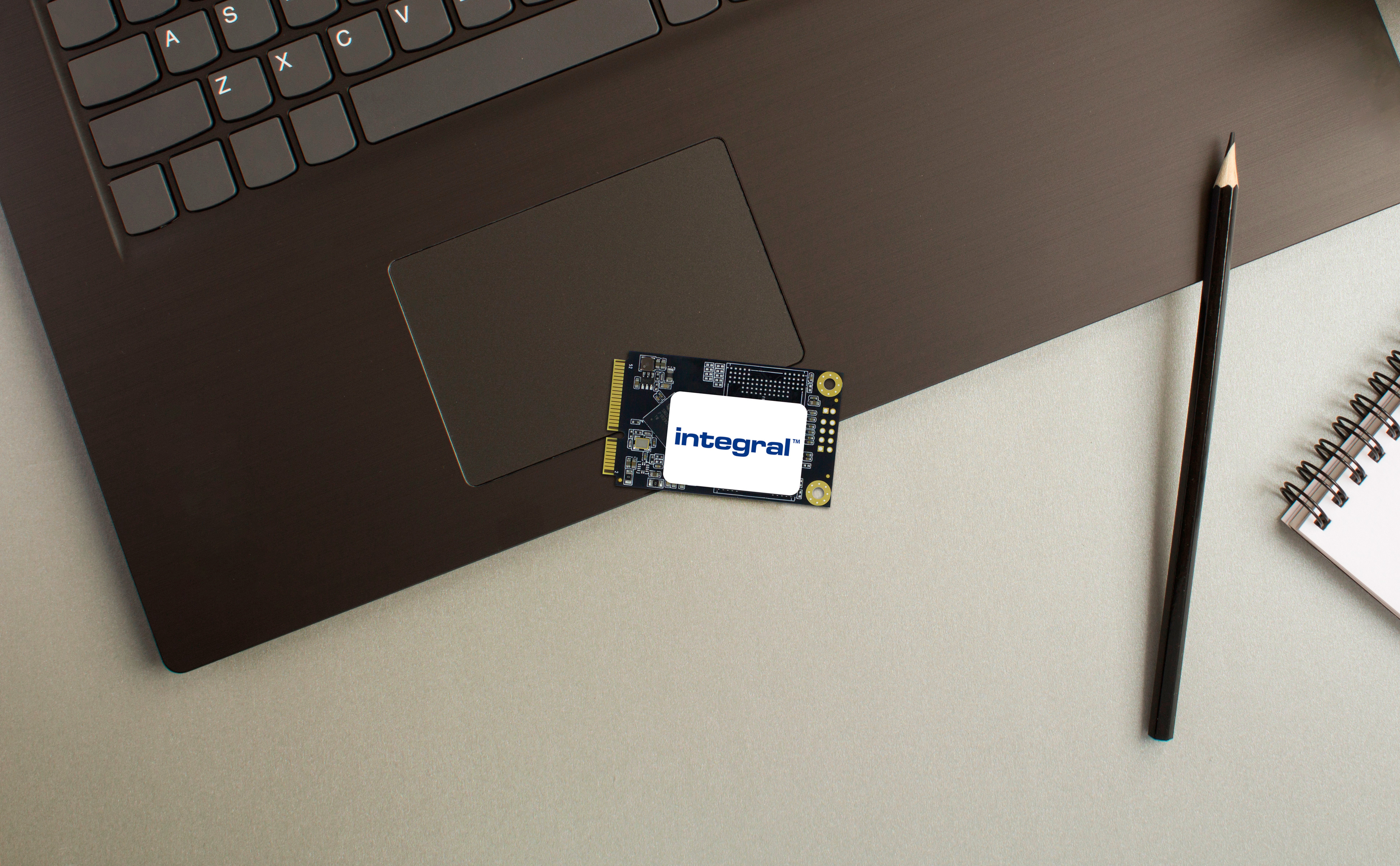 Laptop, Integral SSD, pencil, and notepad on a gray surface. Keyboard keys visible. The background is gray.