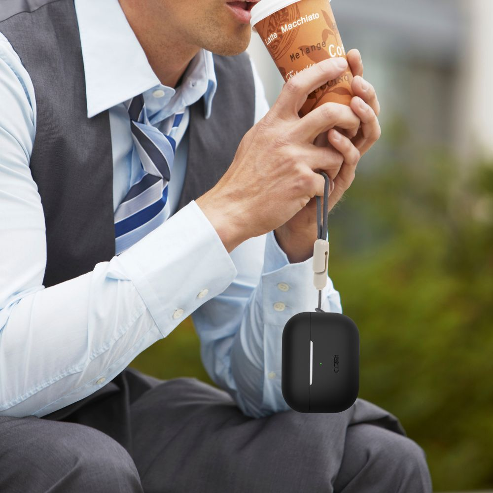 A man in a suit drinks coffee, holding an Airpods case. The background is blurred green. He wears a tie and a vest.
