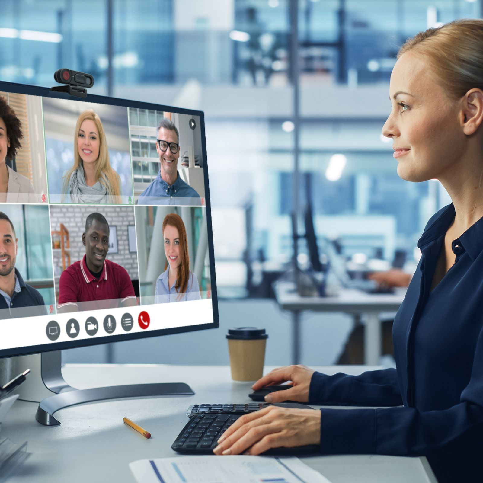 Kobieta w granatowej koszuli podczas rozmowy wideo. Ekran komputera pokazuje innych ludzi. Na stole jest kubek kawy.
