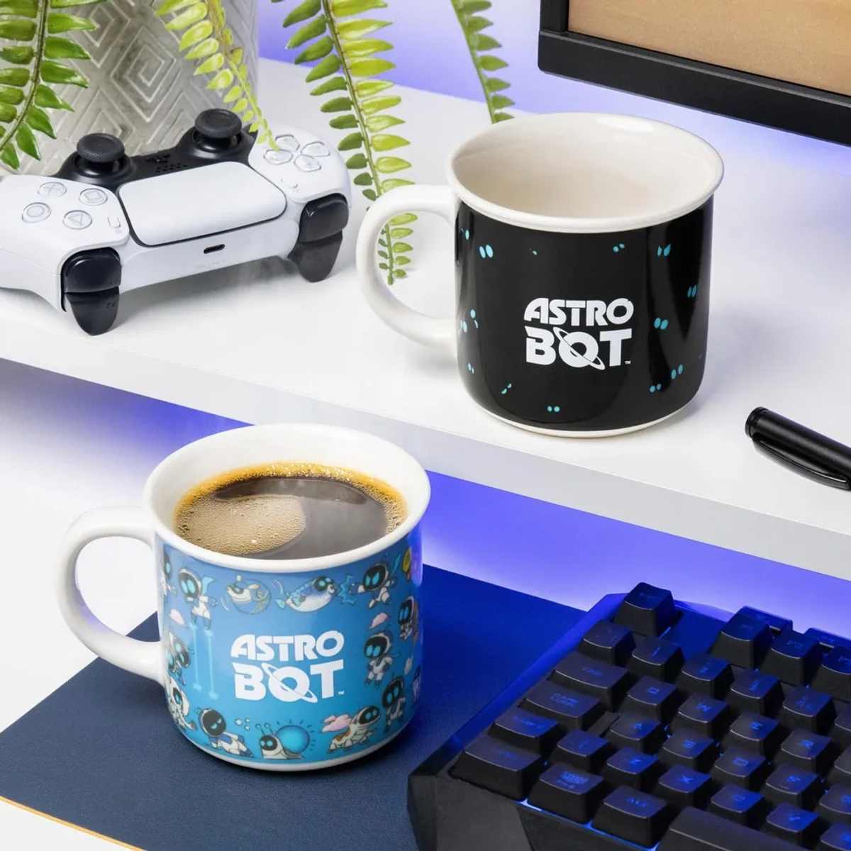 Trzy kubki z logo Astro Bot. Jeden wypełniony kawą, kontroler, klawiatura i roślina na białym biurku.