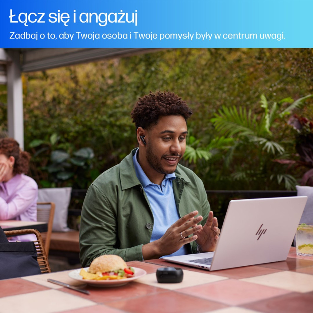 Mężczyzna w słuchawkach prowadzi rozmowę wideo na laptopie. W tle burger, napój i inna osoba.