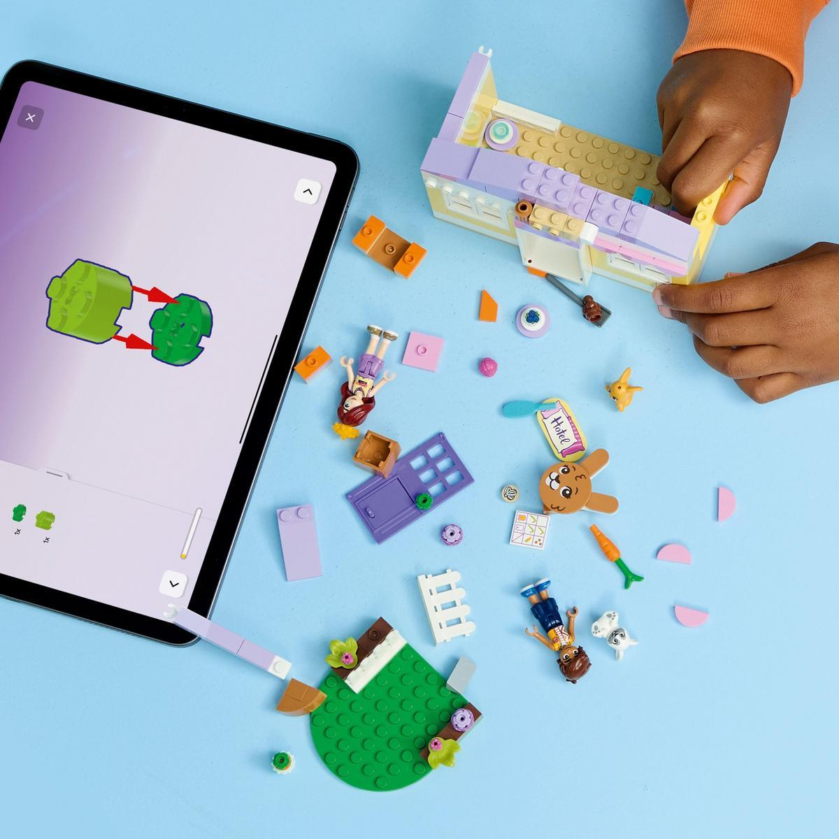 Ręce dziecka w pobliżu klocków Lego. Tablet pokazuje instrukcje. Różne figurki Lego, kwiaty i akcesoria są rozrzucone na niebieskim tle.