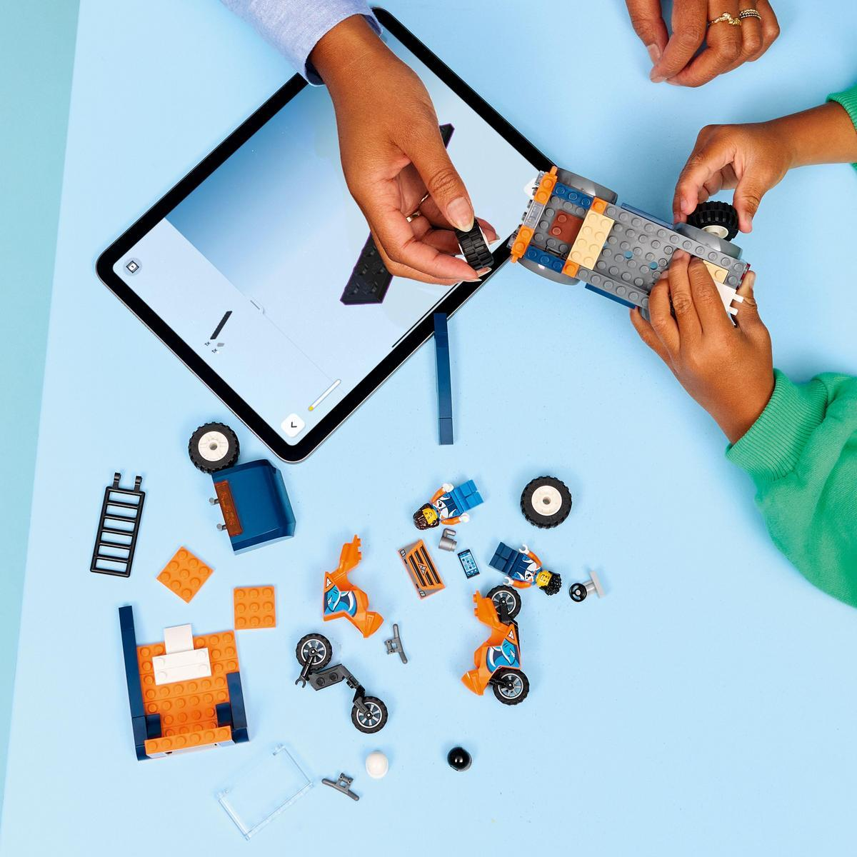 Ręce składające zestaw samochodowy Lego z tabletem pokazującym instrukcje, rozrzucone klocki Lego na niebieskiej powierzchni.
