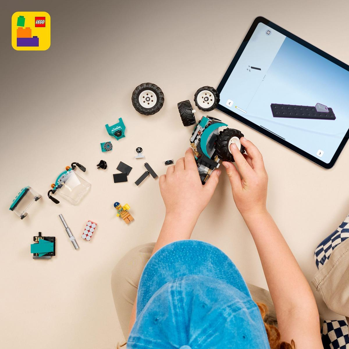 Dziecko składa samochód Lego z instrukcjami na tablecie. Klocki Lego, koła i minifigurka są rozrzucone.