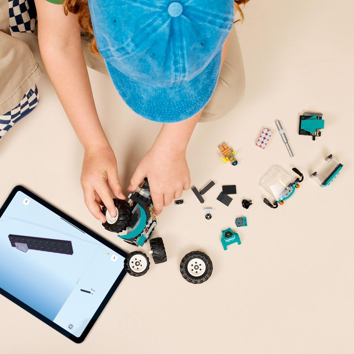 Dziecko w niebieskiej czapce buduje pojazd Lego. Tablet wyświetla instrukcje. Rozrzucone klocki i koła Lego.