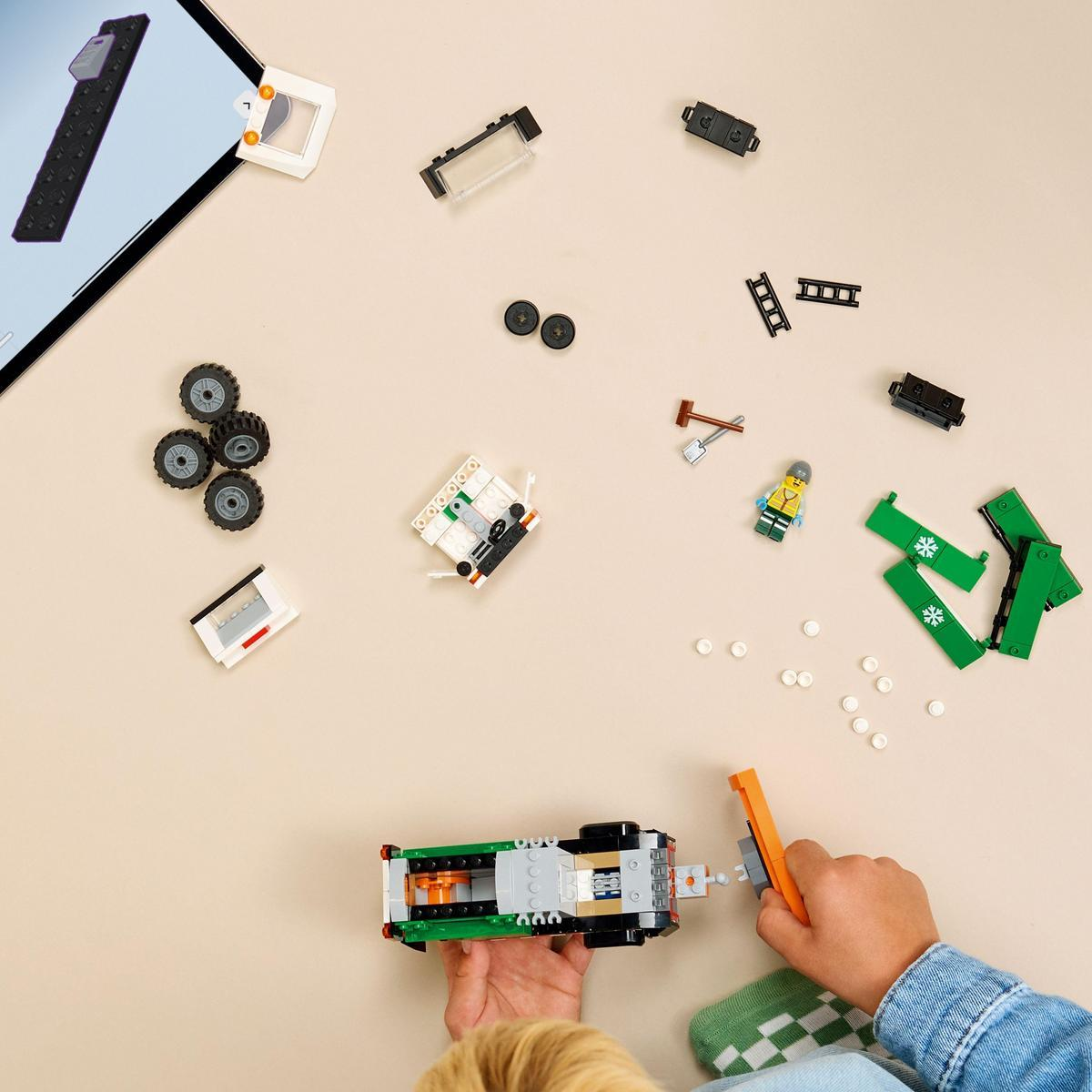 Dziecko składa klocki Lego. Różne elementy są rozrzucone, w tym koła, ciężarówka, figurka i tablet z instrukcjami.