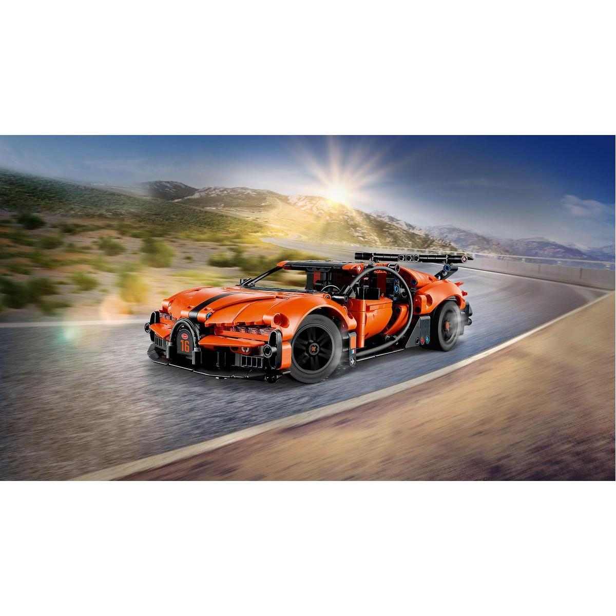 Pomarańczowy samochód sportowy Lego jedzie krętą drogą z górami w tle i jasnym słońcem na niebie.