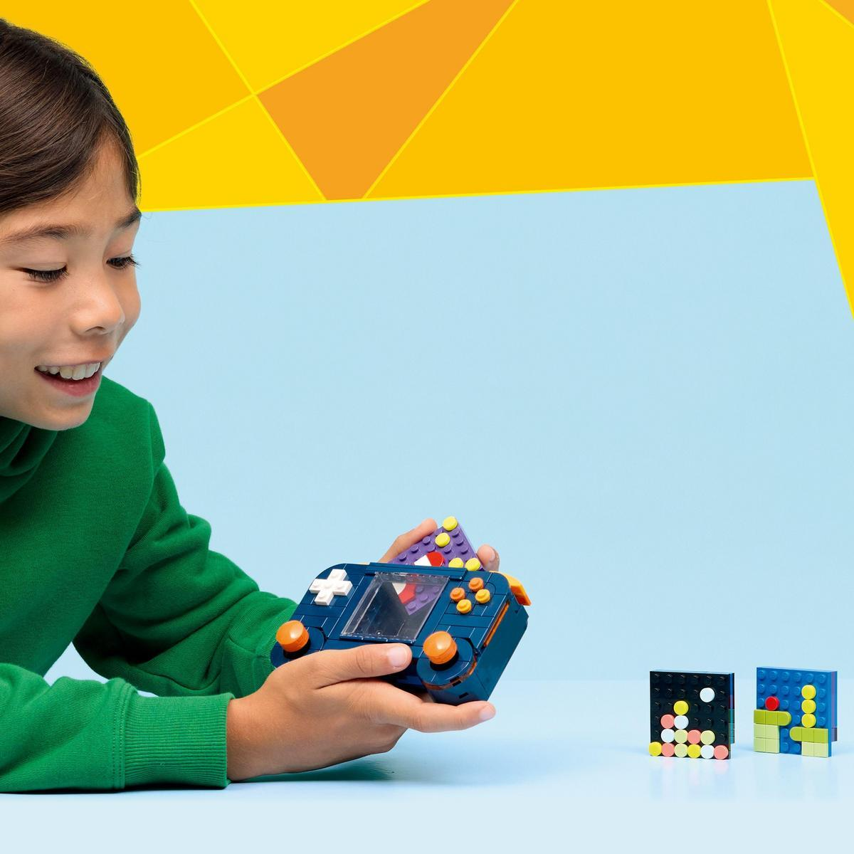 Dziecko trzyma niebieską konsolę do gier wykonaną z klocków. Na stole znajdują się inne figurki z klocków. Tło jest niebieskie i żółte.