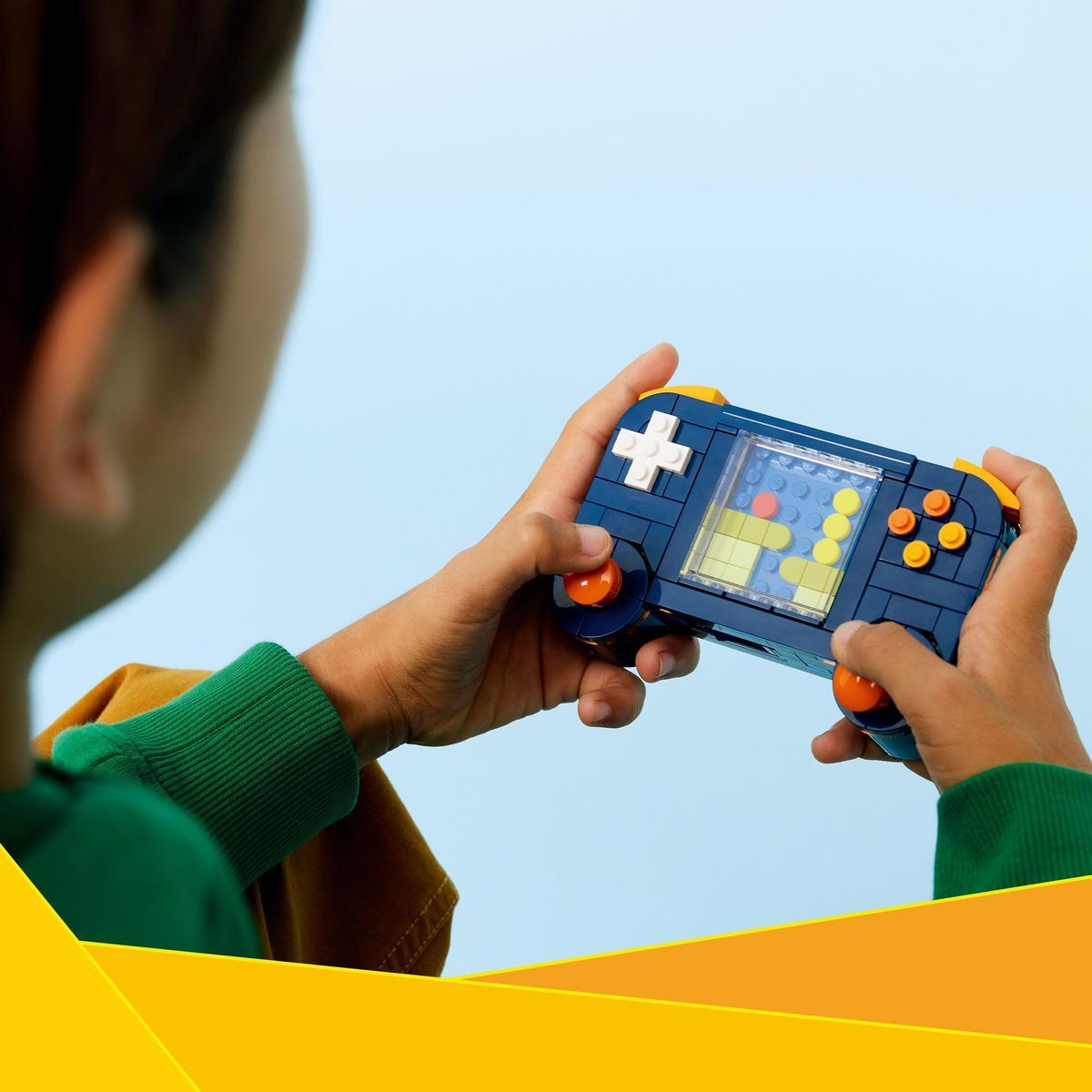 Dziecko trzyma niebieską konsolę do gier Lego. Konsola ma pomarańczowe przyciski i ekran wyświetlający kolorowe klocki. Tło jest jasnoniebieskie.