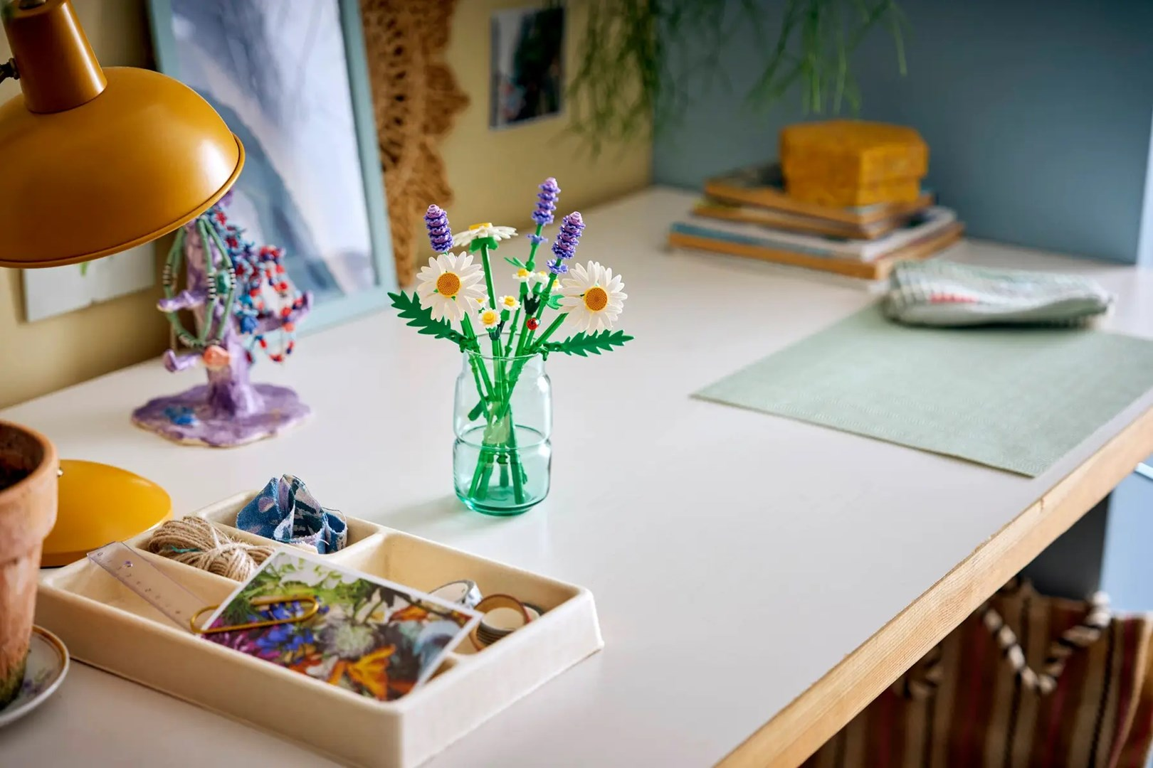 Ein Schreibtisch mit einer Blumenvase, einer Lampe und einem Tablett mit Zubehör. Der Hintergrund umfasst ein Bild und einen Notizblockstapel.