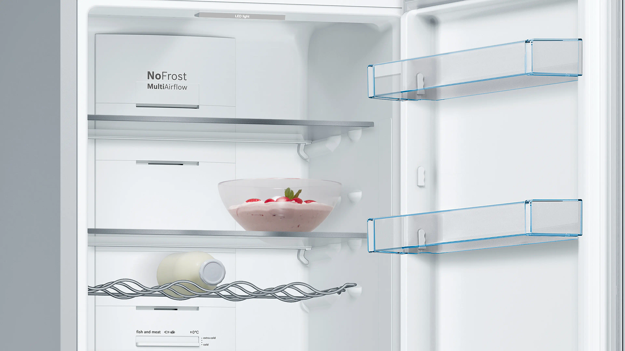 Interno di un frigorifero aperto. È dotato di ripiani, una ciotola di cibo e una bottiglia. Le etichette indicano 'NoFrost' e le impostazioni della temperatura.