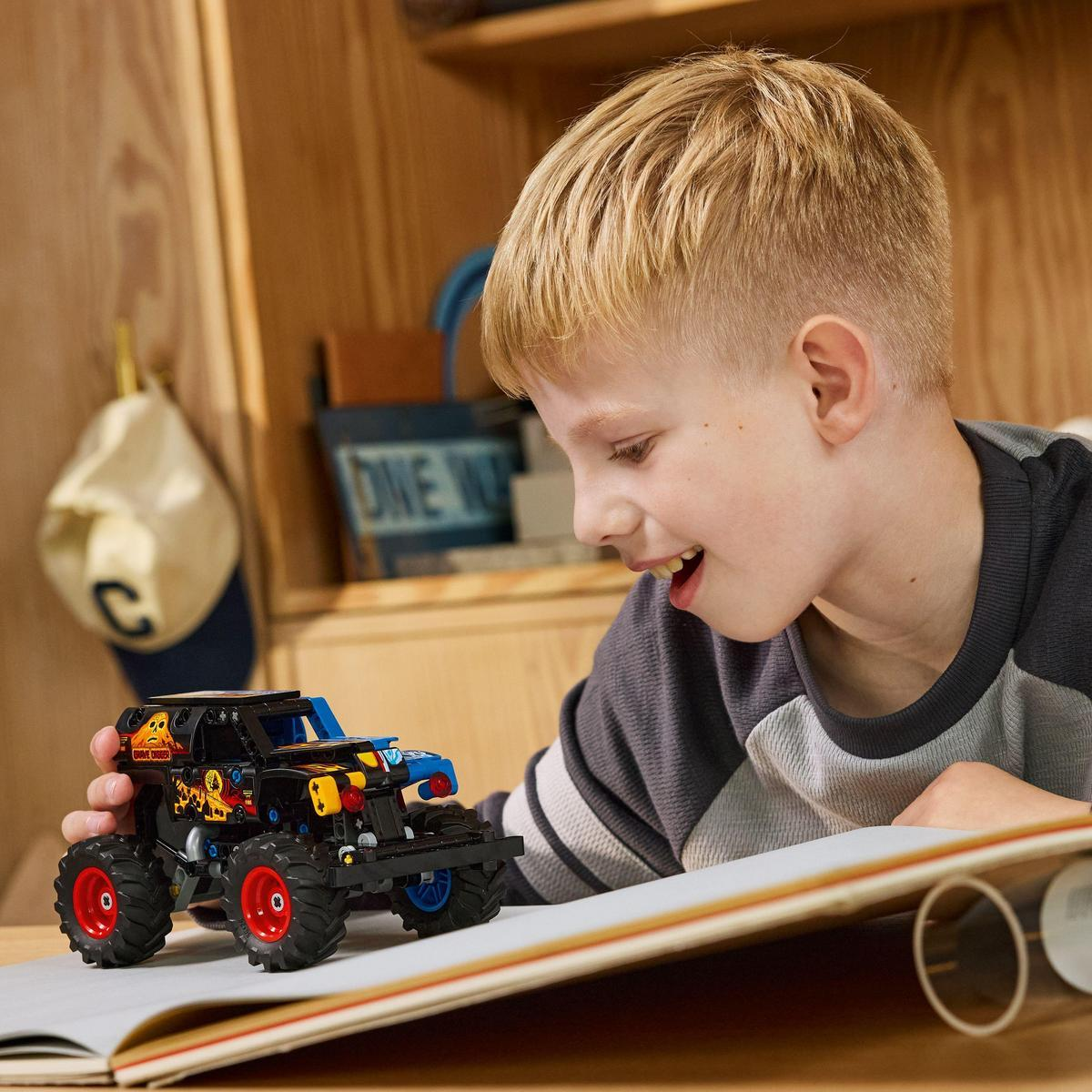 Chłopiec uśmiecha się do czarno-niebieskiej zabawki ciężarówki. Ma czerwone koła. Chłopiec czyta książkę na drewnianej powierzchni.