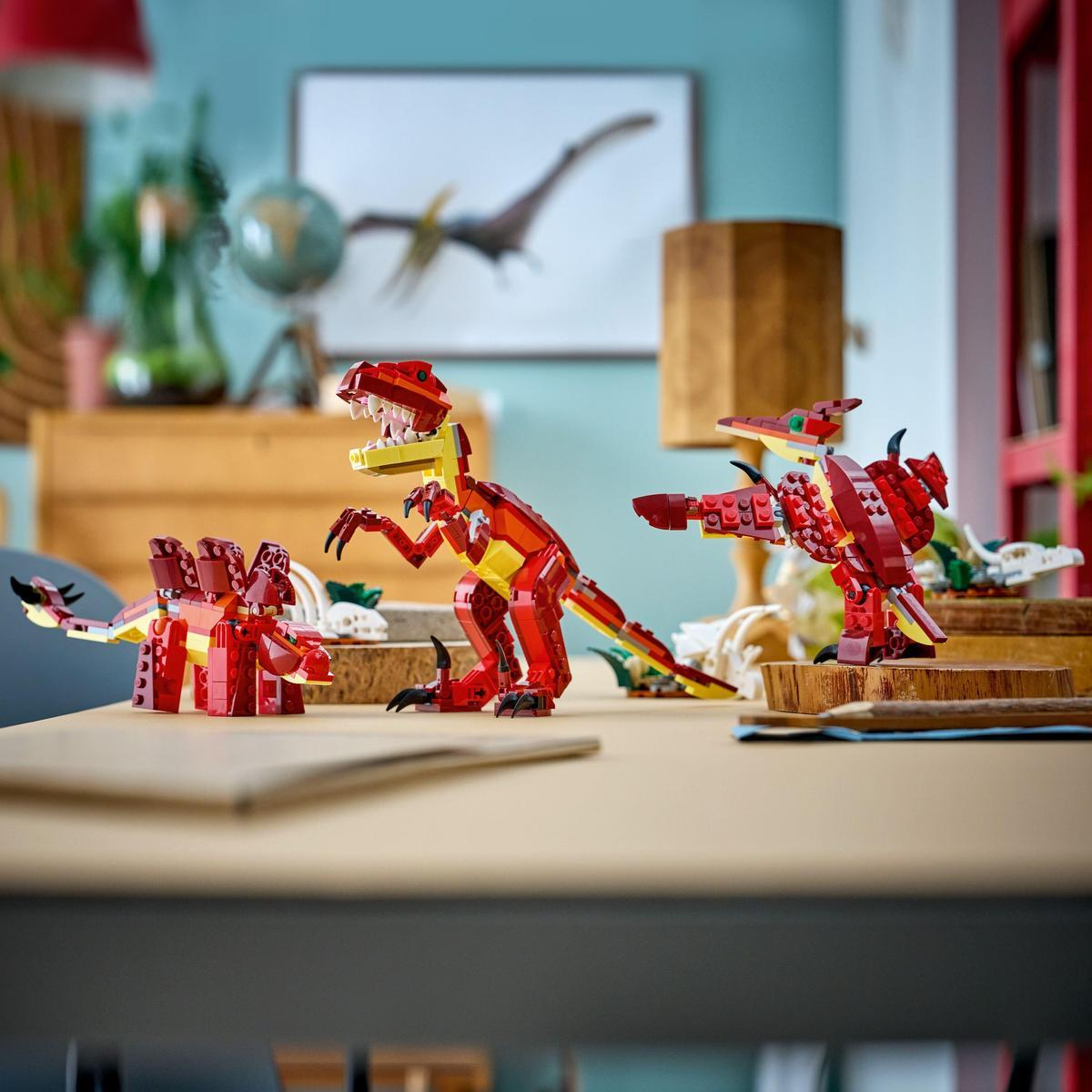 Dinozaury LEGO na biurku. Czerwone i żółte modele na pierwszym planie. Oprawiony obraz pterodaktyla i inne dekoracje w tle.