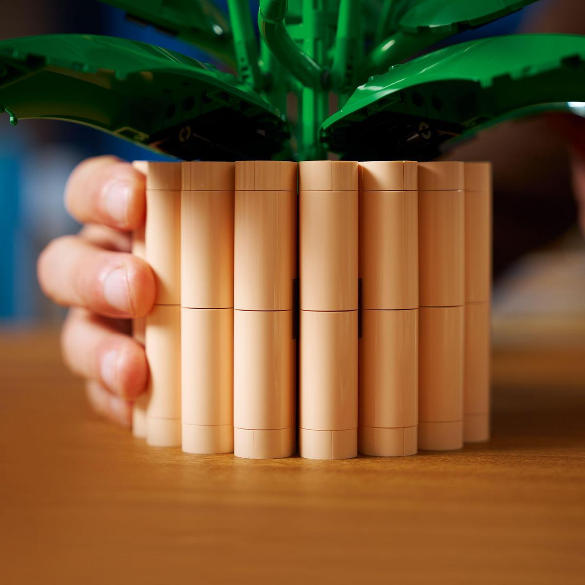 Ręka trzyma beżową doniczkę z zielonymi plastikowymi liśćmi. Doniczka wykonana jest z pionowych walców. Tło to rozmyte drewno i niebieski.