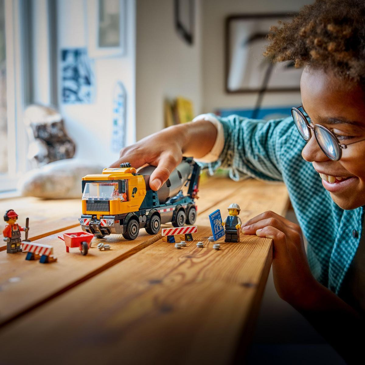 Dziecko w okularach bawi się ciężarówką do betonu Lego. Figurki Lego i bariery budowlane leżą na drewnianej powierzchni.