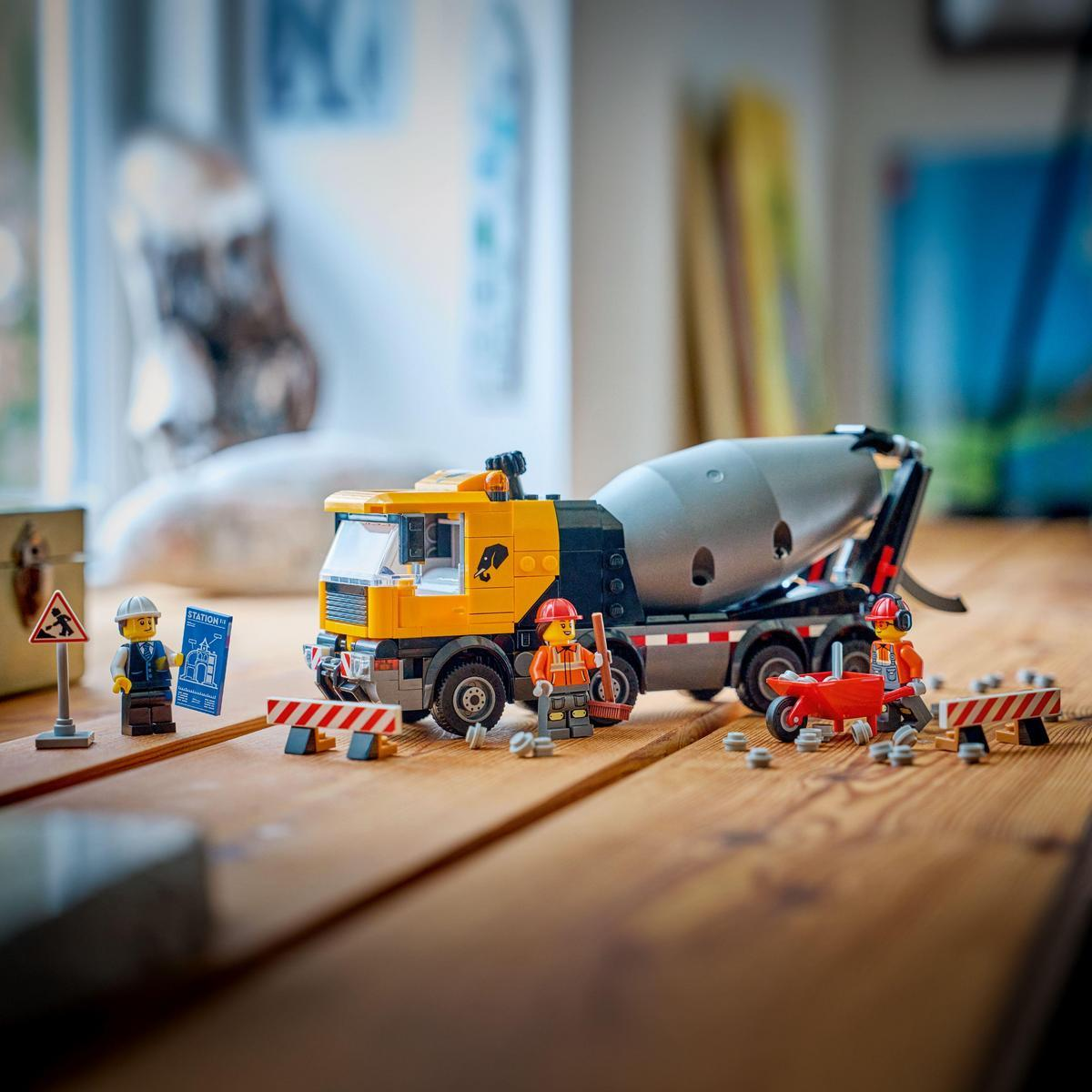 Scena budowy Lego. Żółto-szara betoniarka, robotnicy, czerwony wózek i bariery drogowe na drewnianej powierzchni.