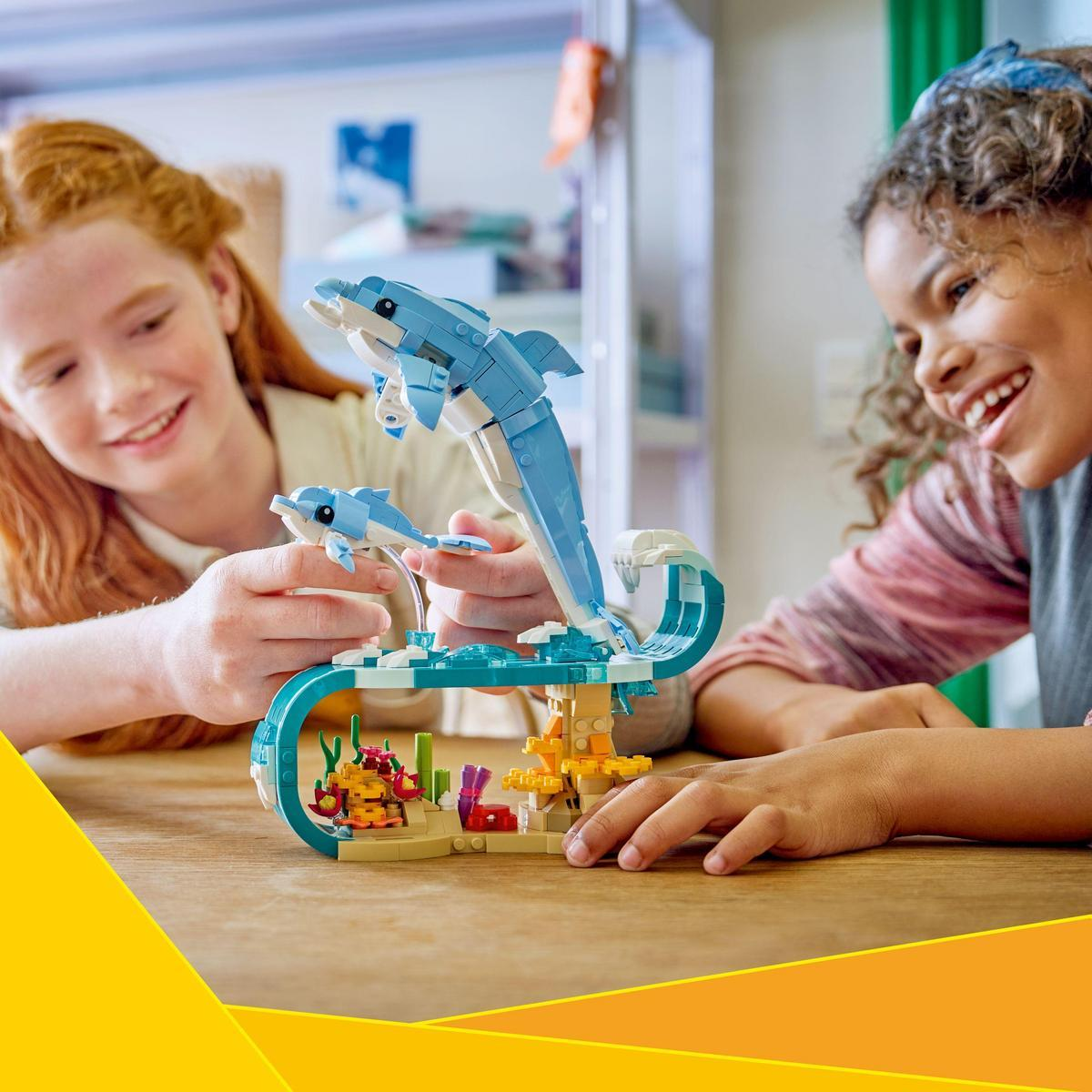 Dwie dziewczynki uśmiechają się podczas budowania zestawu Lego z delfinami. Zestaw przedstawia niebieskie delfiny skaczące nad sceną oceanu.