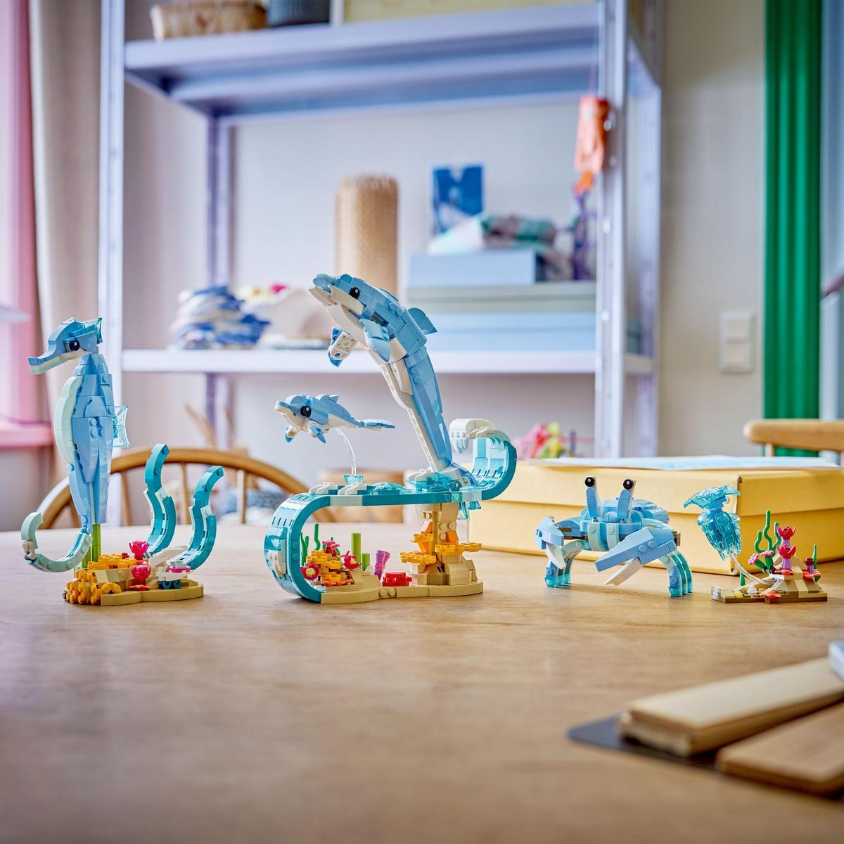 Stół prezentuje morskie stworzenia Lego: konik morski, rodzina delfinów, krab i inne elementy morskie. Tło przedstawia półki i okno.