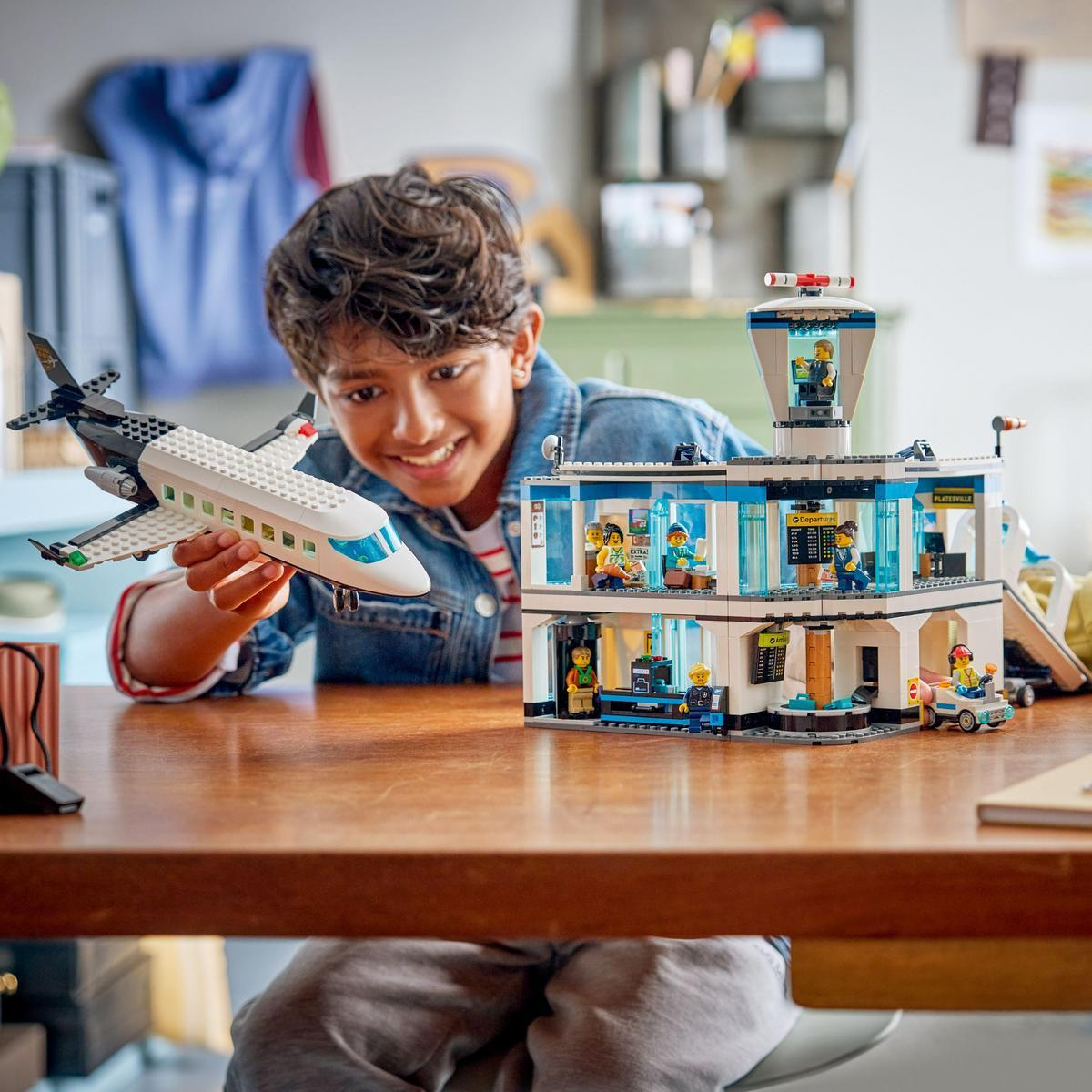 Chłopiec trzyma zabawkę samolotu. Zestaw lotniska Lego stoi na stole. Chłopiec się uśmiecha. Na lotnisku są ludzie w środku.