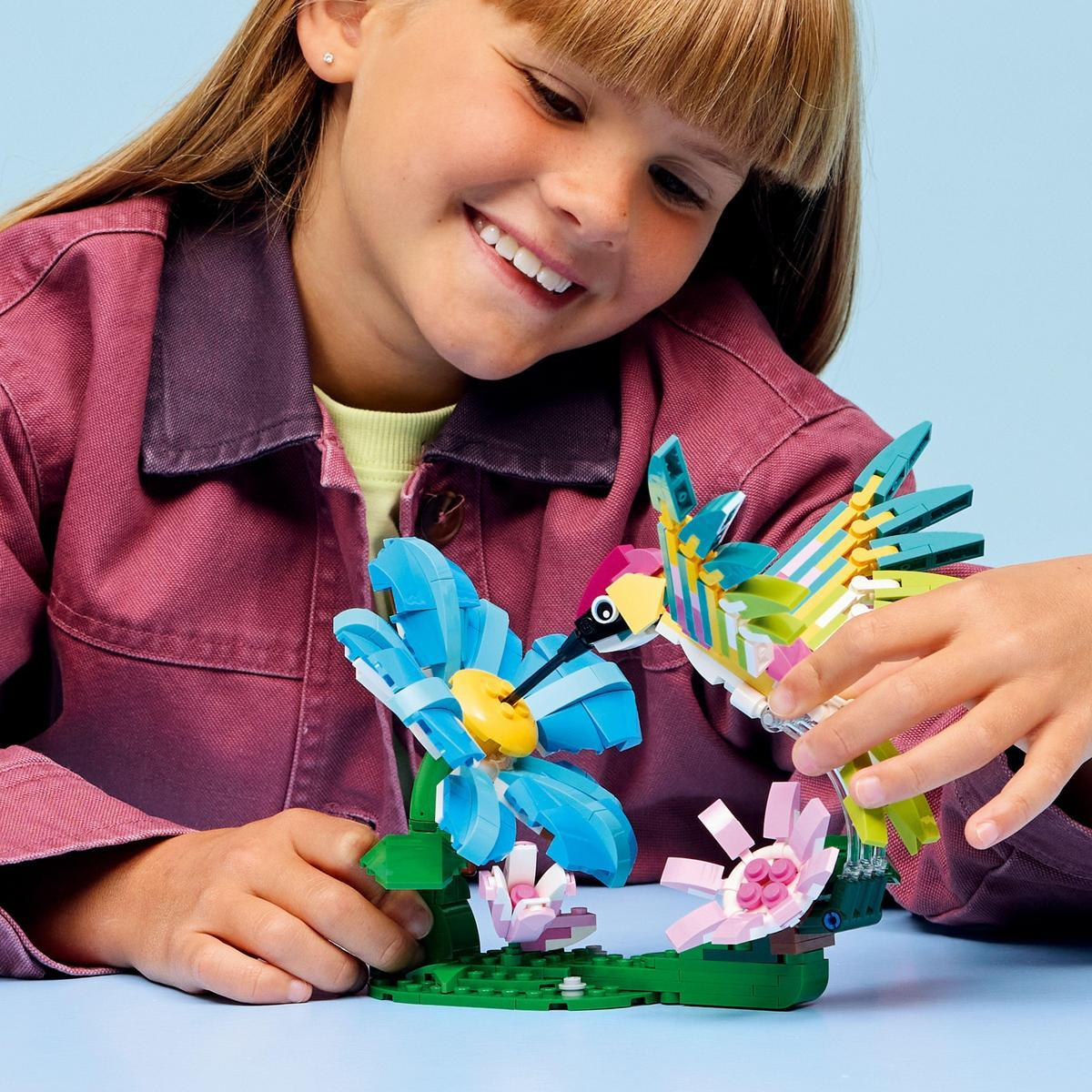 Dziewczyna uśmiecha się do kolibra i kwiatów LEGO. Ptak jest kolorowy. Kwiaty są niebieskie i różowe. Scena jest na niebieskim tle.