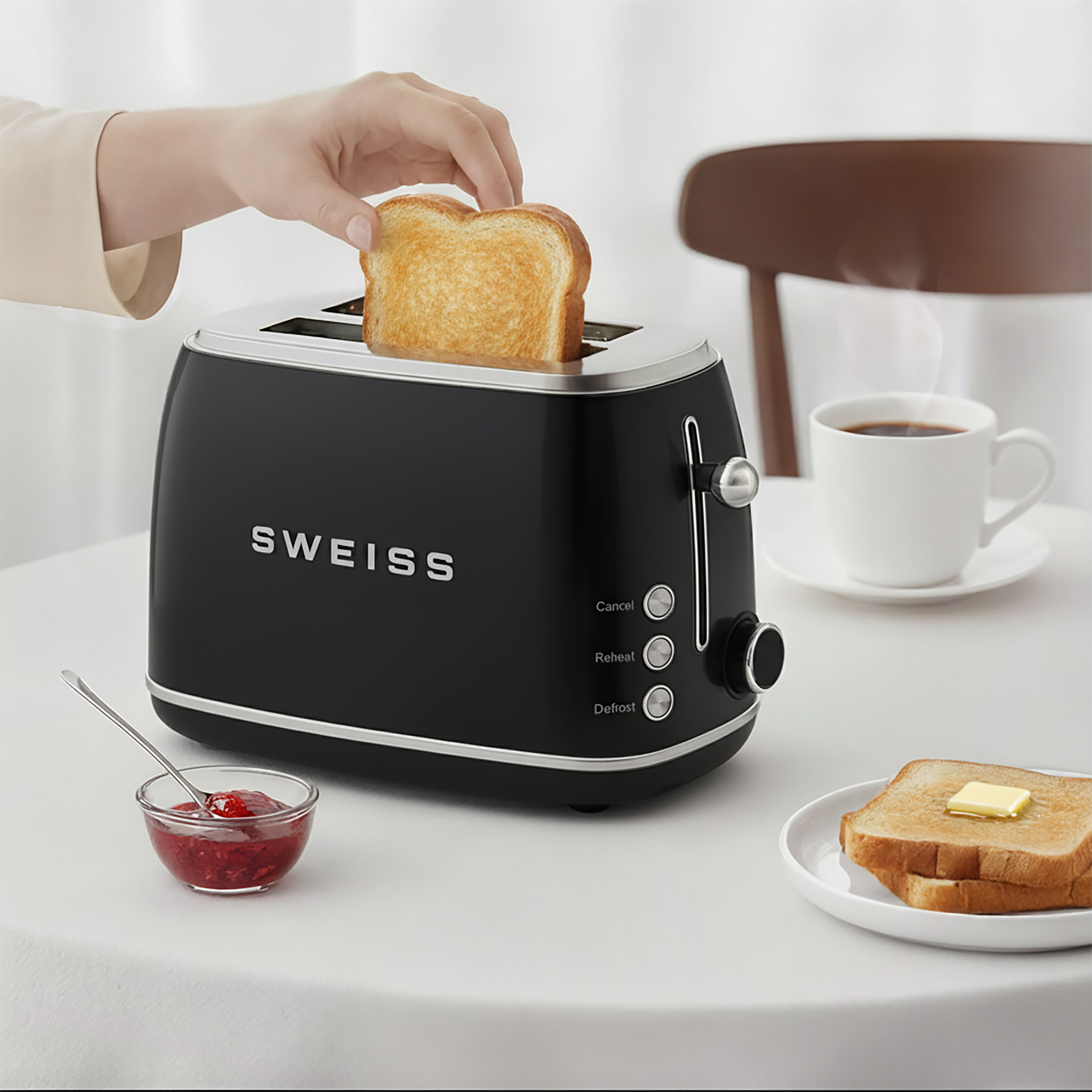 Eine Hand legt Toast in einen schwarzen Toaster. Eine Tasse Kaffee, Marmelade und Toast mit Butter sind ebenfalls auf dem Tisch.