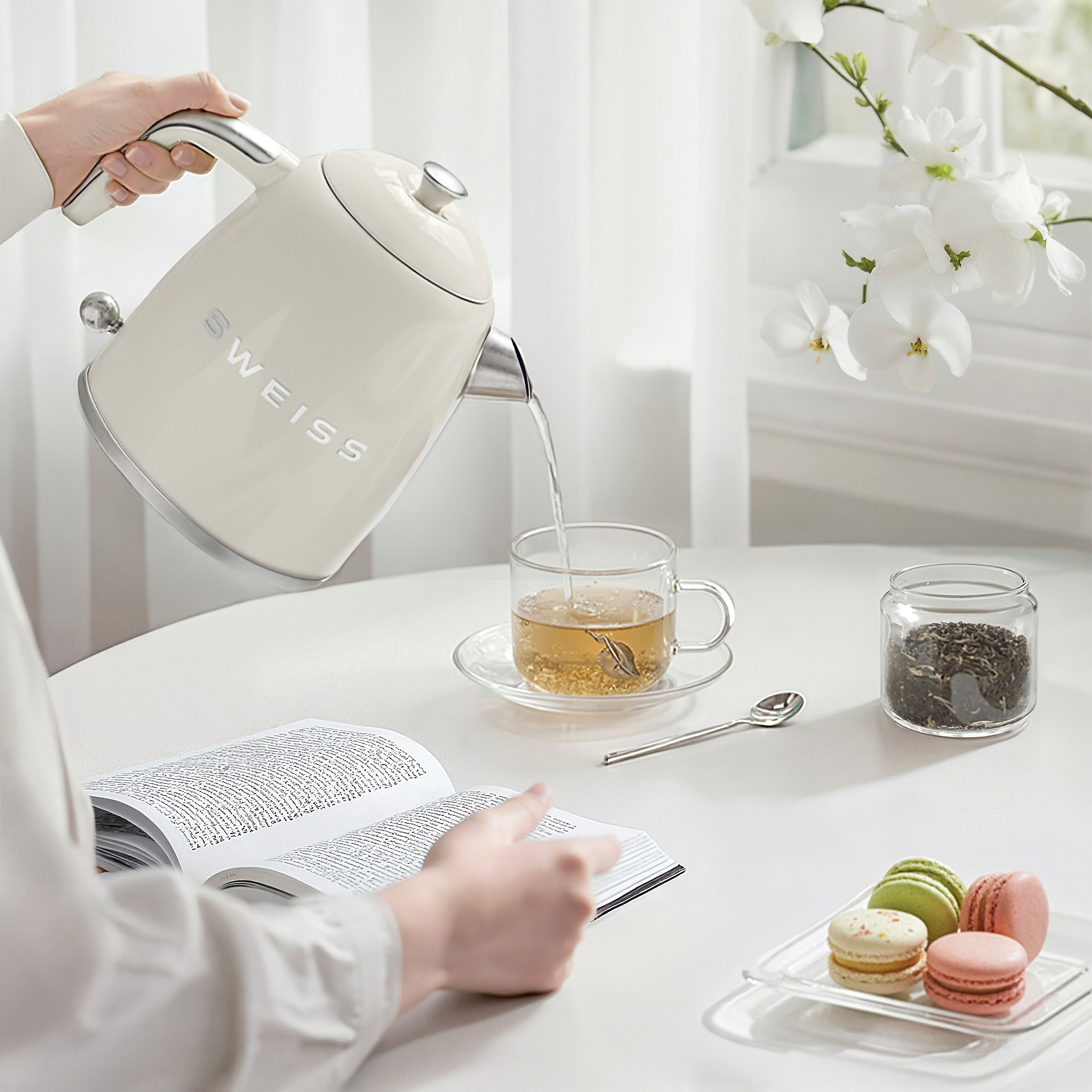 Eine Person gießt heißes Wasser aus einem cremefarbenen Wasserkocher in eine Teetasse. Makronen und ein Buch liegen auf dem Tisch.