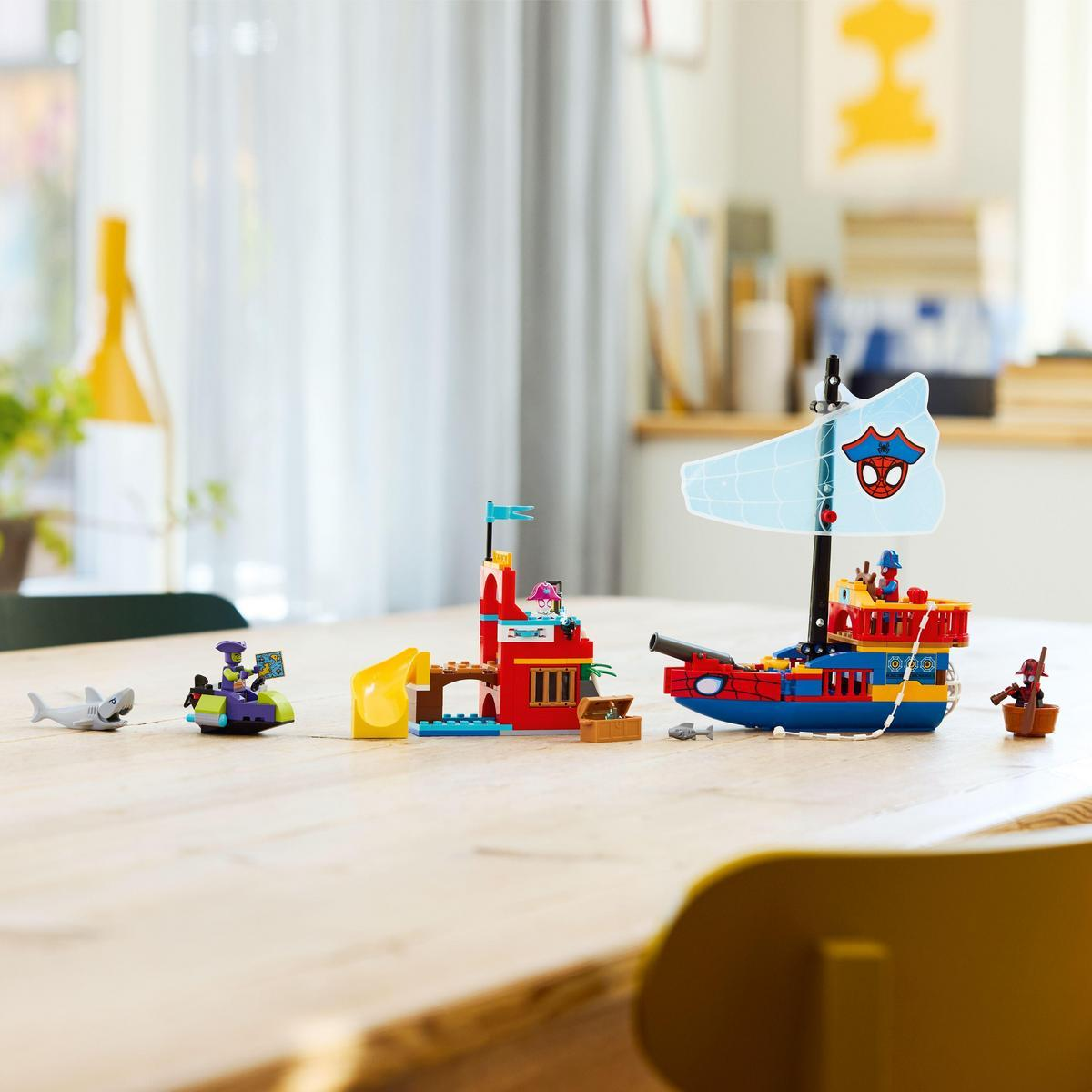 Zestaw Lego na stole. Łódź Spider-Mana z żaglem i armatą, czerwony budynek ze zjeżdżalnią, figurki i rekin.