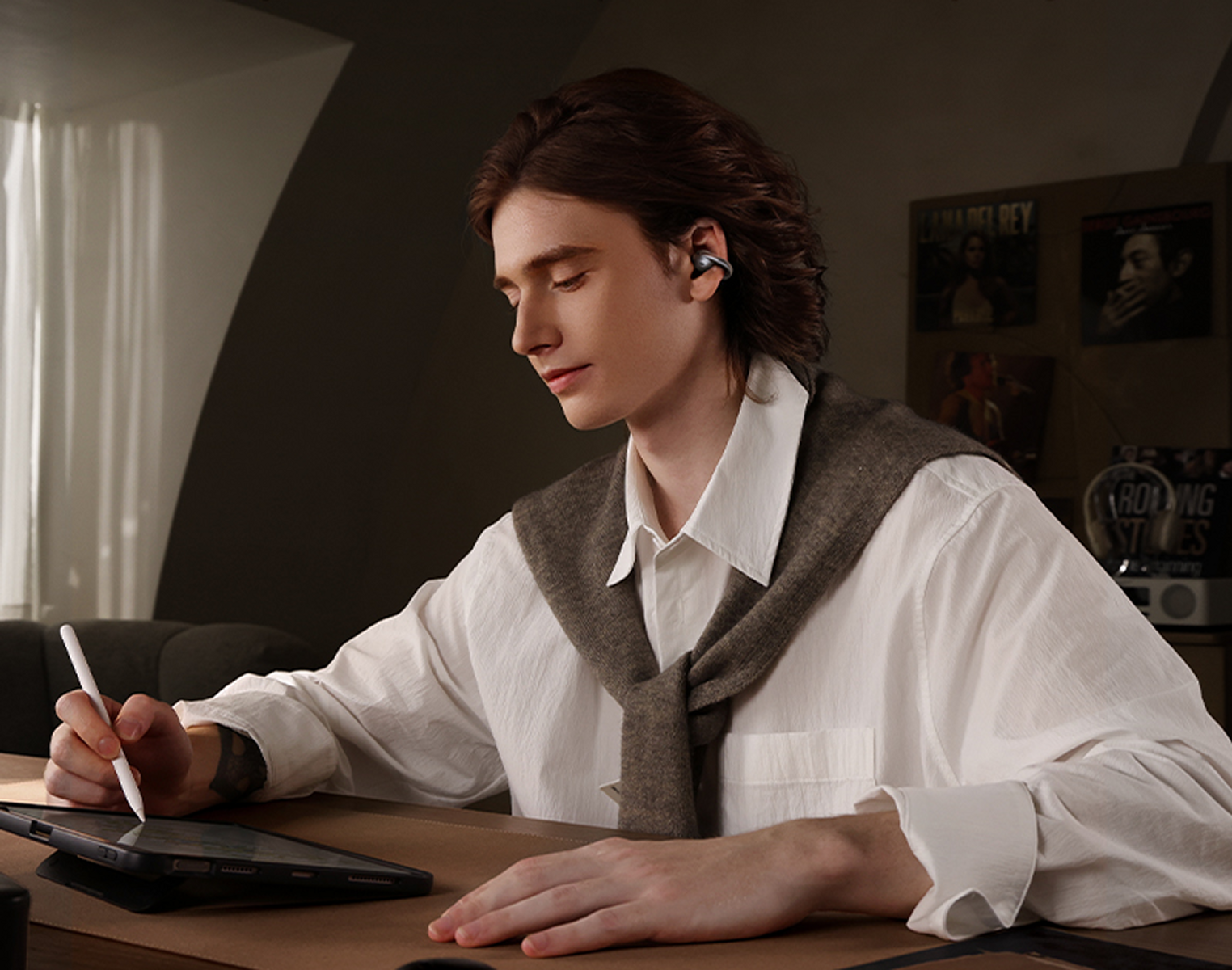 Un uomo con auricolari disegna su un tablet con una penna. Indossa una camicia bianca e un maglione. Sfondo: poster.
