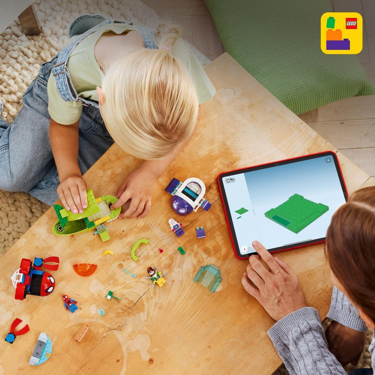 Dziecko i dorosły budują Lego na drewnianym stole, kierując się tabletem. Klocki i figurki Lego są rozrzucone. Zielona poduszka w tle.