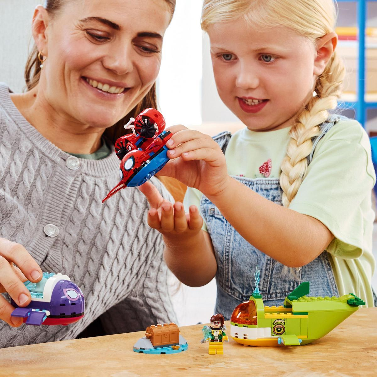 Kobieta i dziewczynka bawią się zabawkami samolotami i figurkami. Dziewczynka trzyma czerwono-niebieski samolot. Inne zabawki na stole.