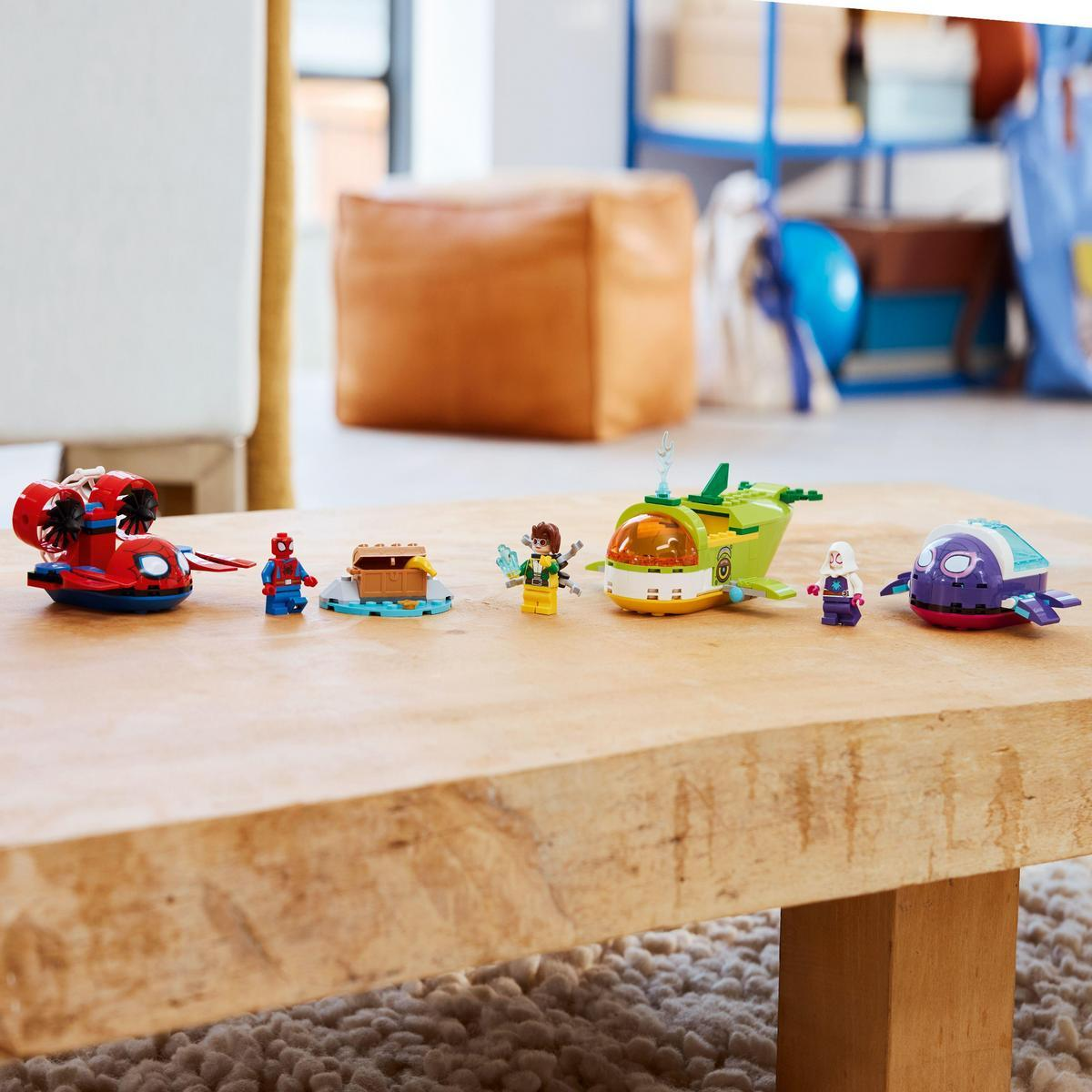 Zestaw Lego na drewnianym stole. Zawiera figurki superbohaterów, statki i skrzynię. Tło przedstawia pokój z brązową poduszką.