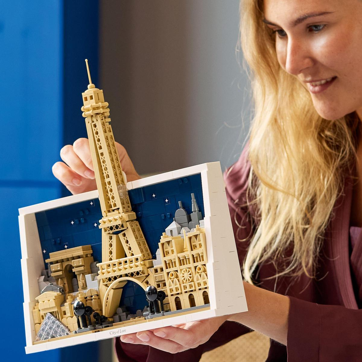 Kobieta trzyma dioramę Lego Paryża z Wieżą Eiffla. Tło to niebieskie nocne niebo.