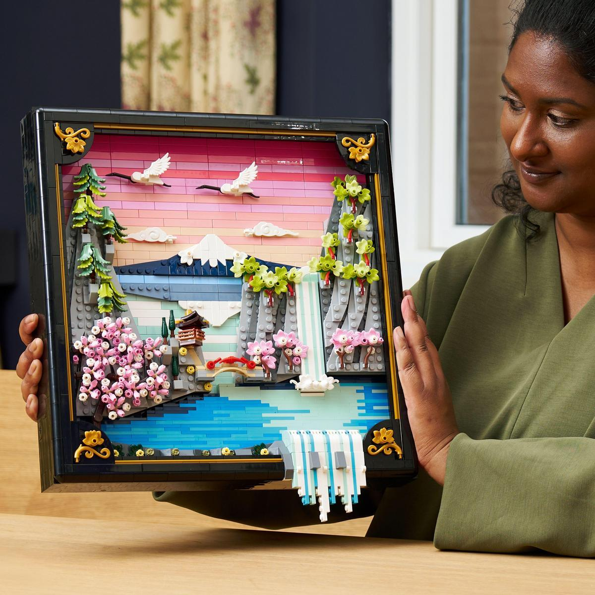 Kobieta trzyma dioramę Lego przedstawiającą japoński krajobraz, w tym górę, jezioro, kwiaty wiśni i wodospad z latającymi ptakami.
