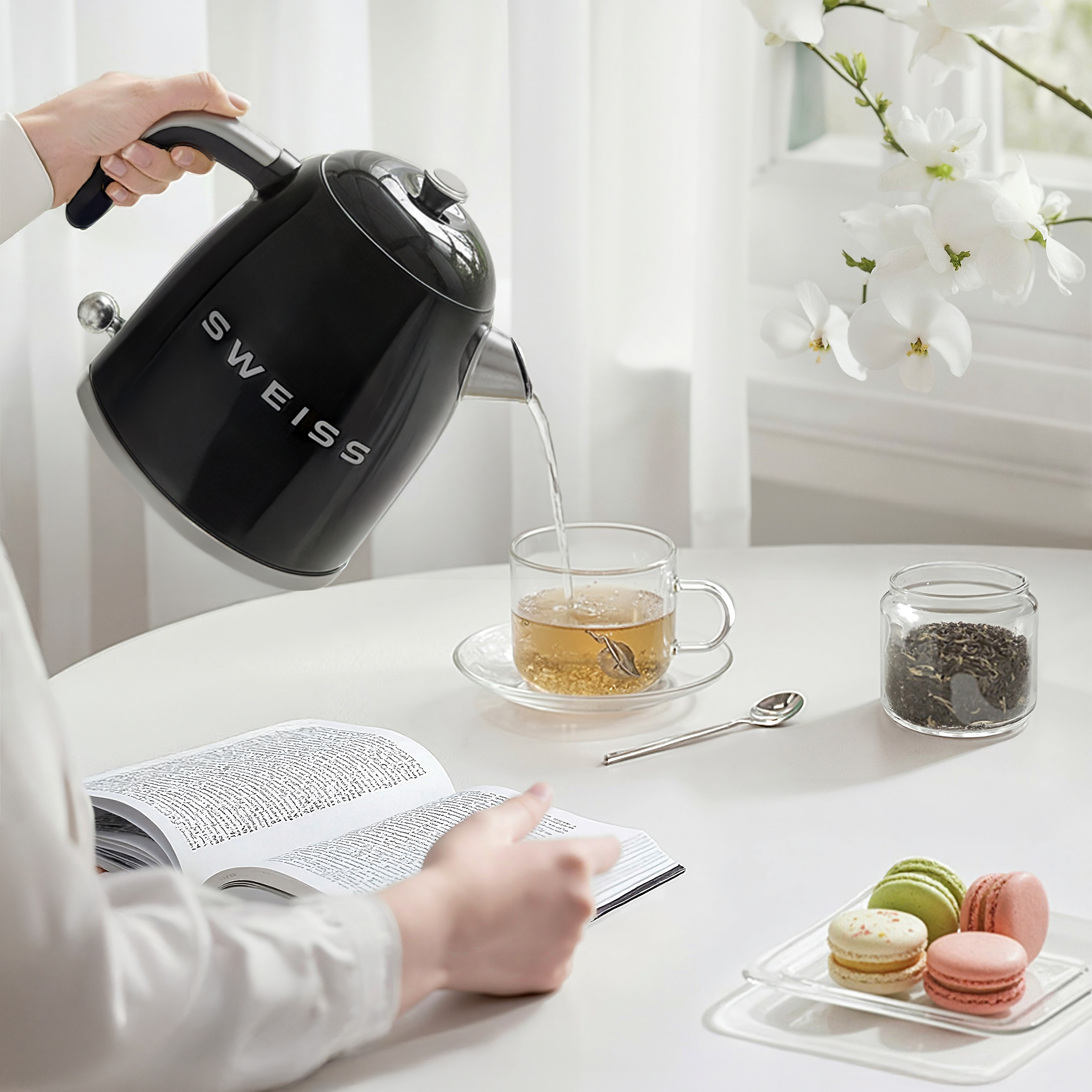 Eine Person gießt Wasser aus einem schwarzen Wasserkocher in ein Glas Tee. Makronen, Teeblätter, ein Buch und Blumen liegen auf dem Tisch.