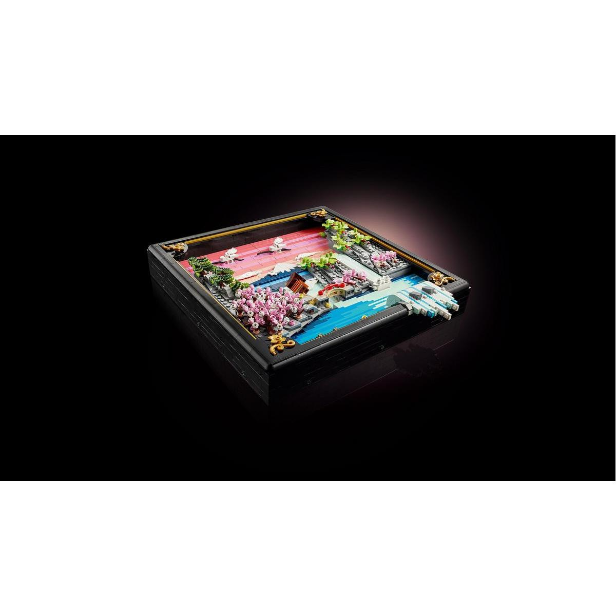 Miniaturowa scena japońskiego ogrodu w czarnej ramie, z mostem, różowymi drzewami, wodą i żywym zachodem słońca.