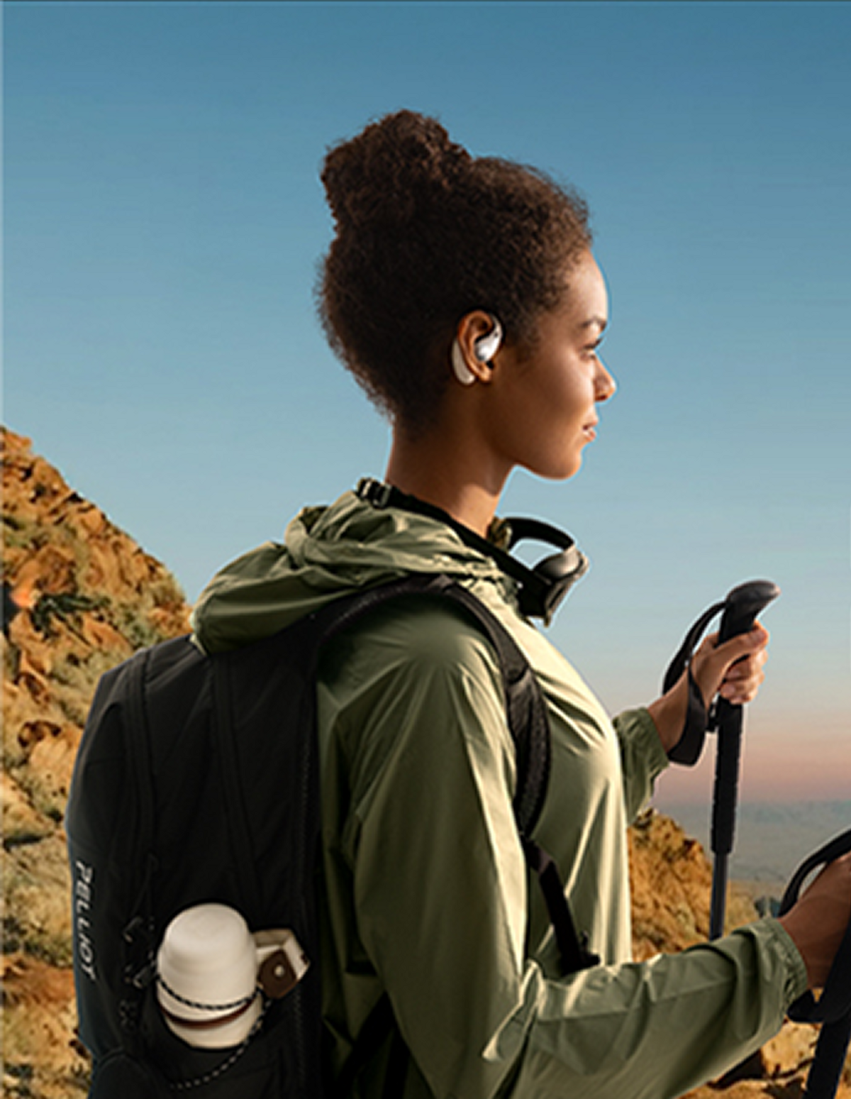Eine Frau wandert mit Ohrhörern, Rucksack und Trekkingstöcken, Berge im Hintergrund, trägt eine grüne Jacke.