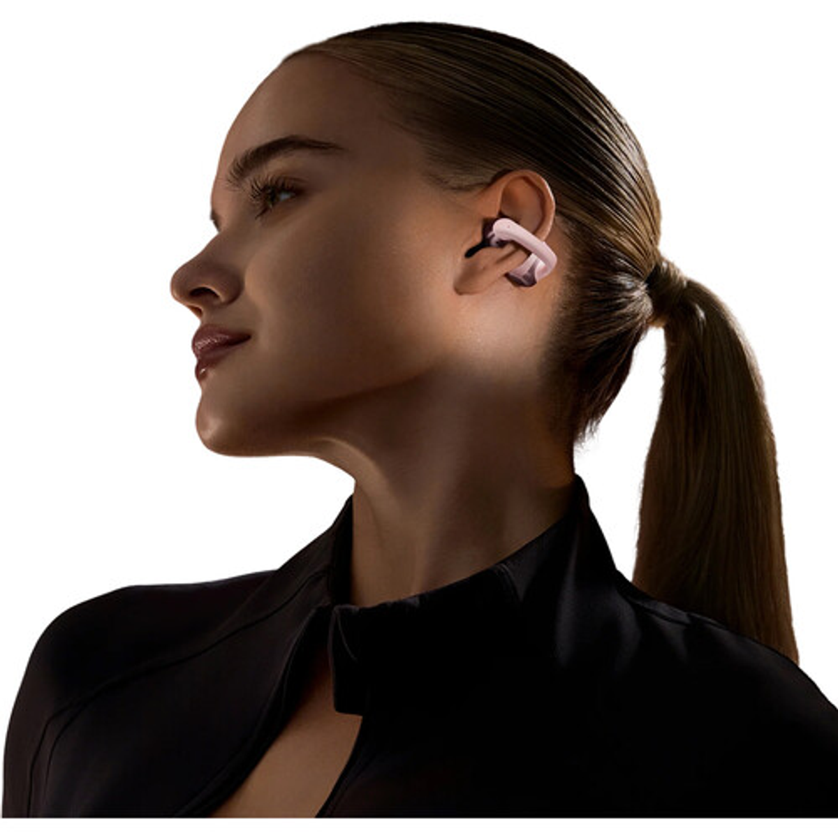 Kobieta w różowo-czarnych słuchawkach, zwrócona w lewo. Ma ciemne włosy w kucyku, czarną kurtkę, tło jest białe.