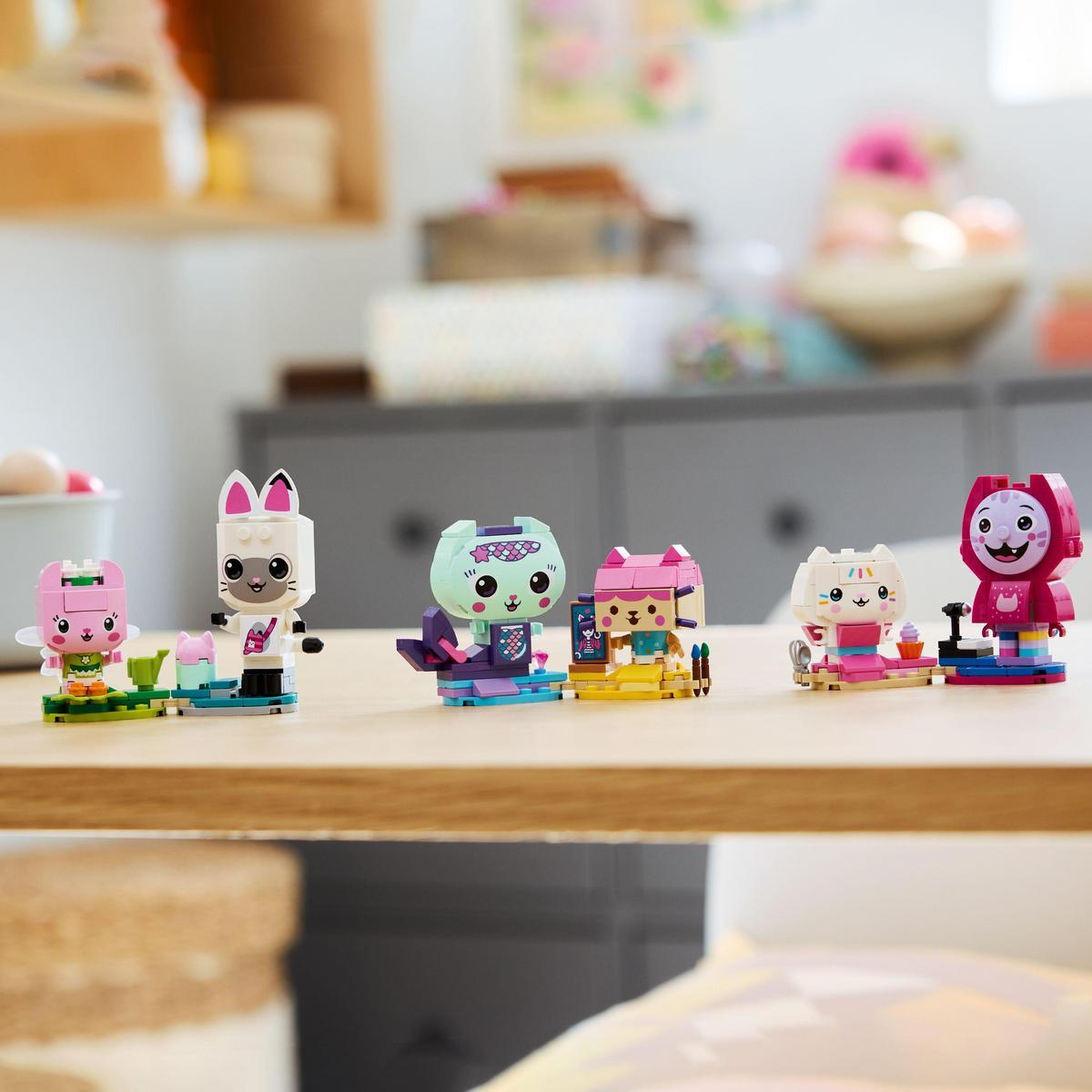 Pięć figurek kota Lego na drewnianym stole. Tło przedstawia rozmyty pokój z półkami i szafką.