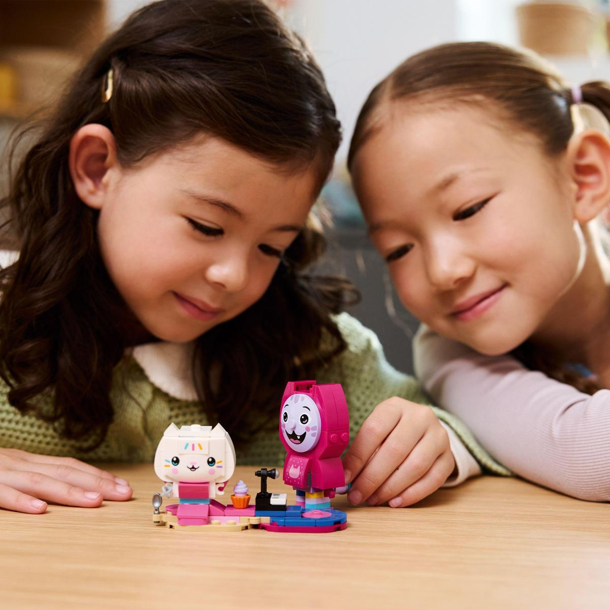 Dwie młode dziewczyny patrzą na kolorowe figurki Lego kota i różowej postaci na drewnianym stole.