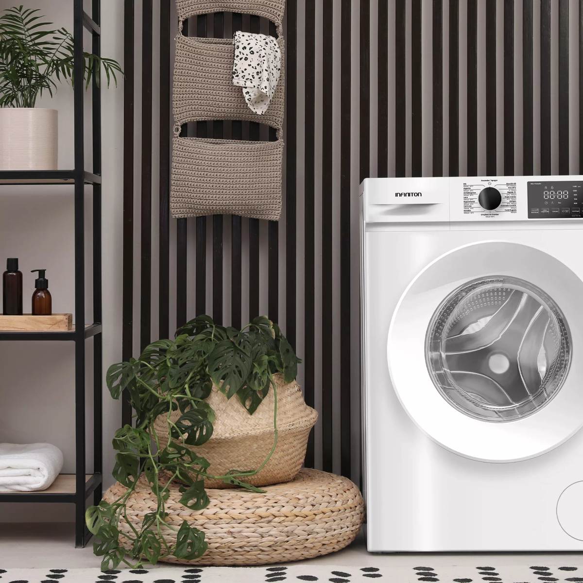 Una lavadora blanca está junto a una cesta con una planta. Hay una pared decorativa detrás.