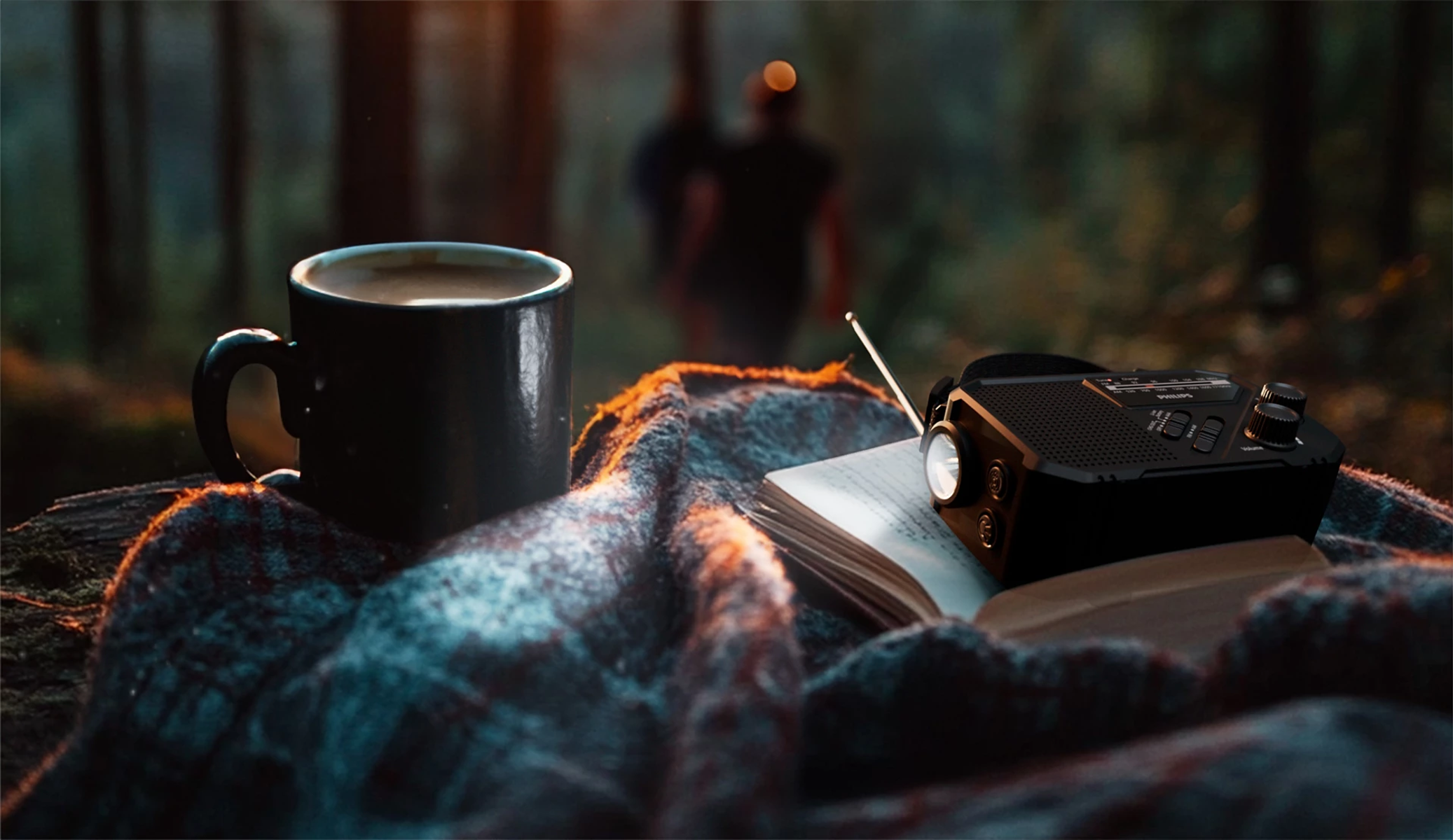 Kubek kawy, radio i książka leżą na kocu. Dwie postacie idą w rozmytym tle lasu.