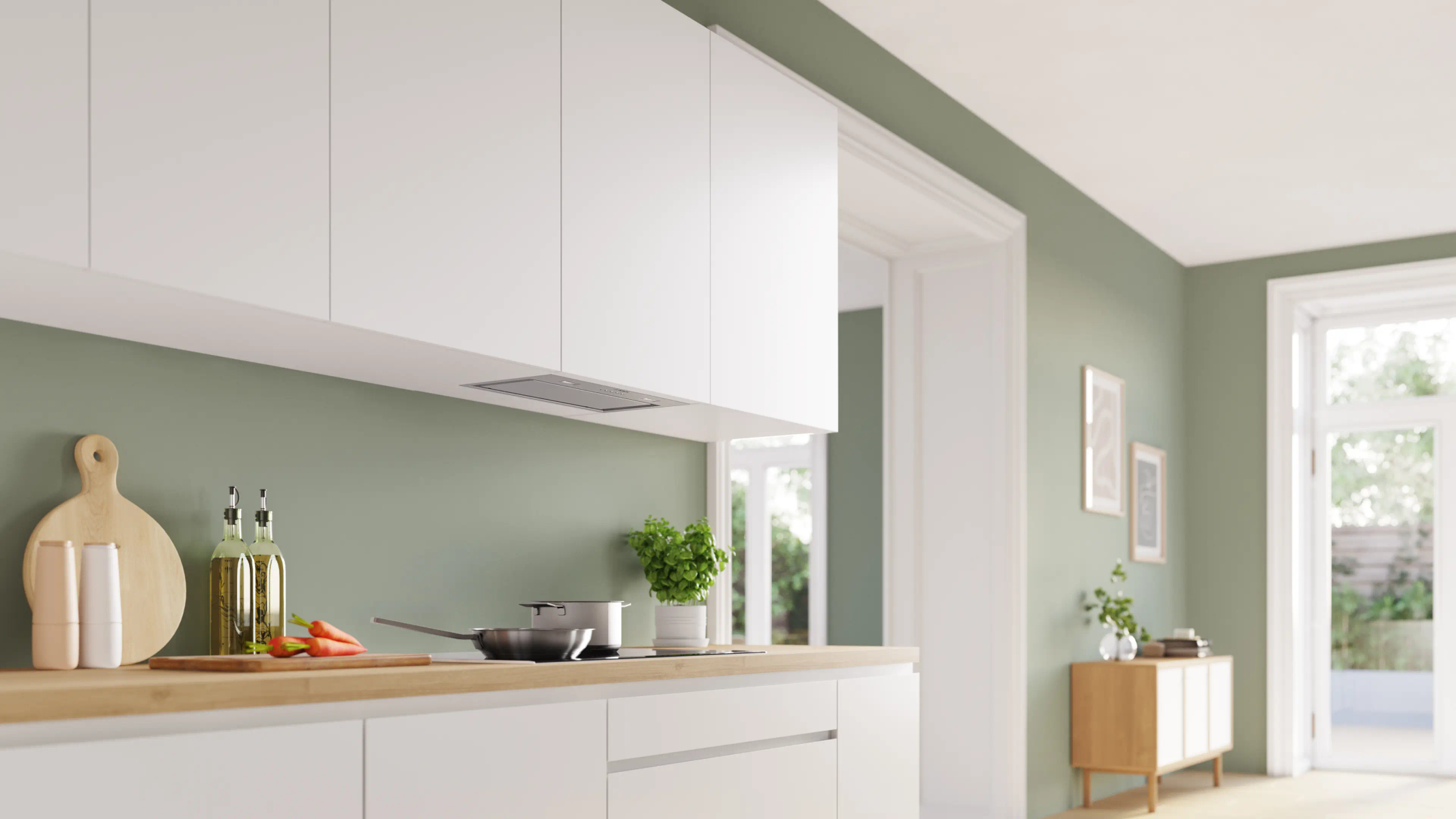 Una cucina moderna con mobili bianchi, piano di lavoro in legno e parete verde salvia. Carote, bottiglie d'olio e una padella sono sul bancone.
