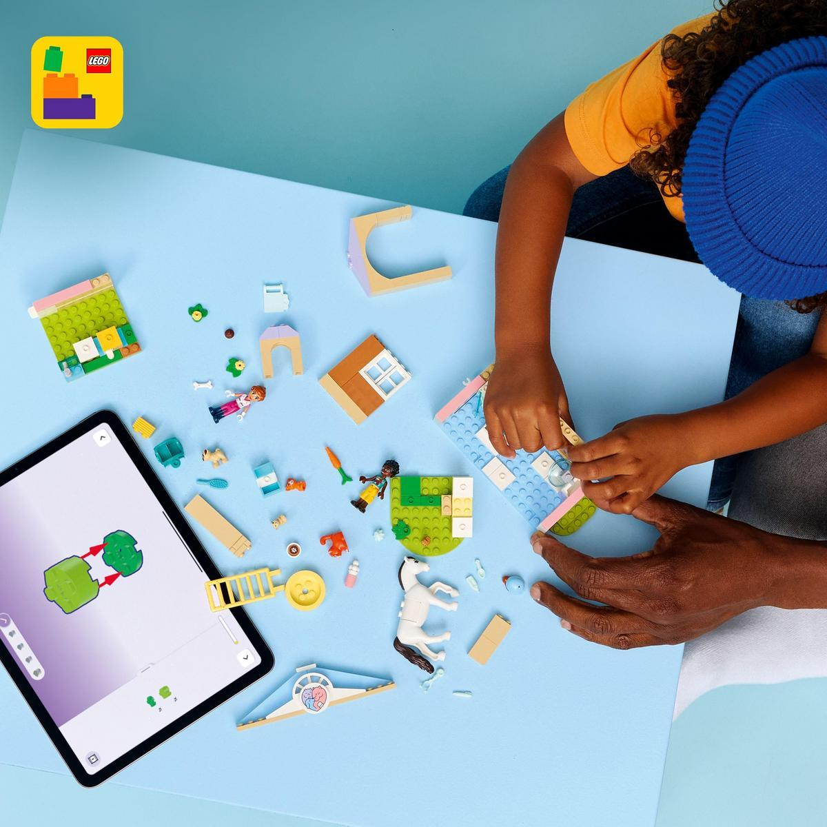 Ręce składające zestaw Lego na niebieskiej powierzchni. Tablet pokazuje instrukcje. Rozrzucone klocki Lego, w tym koń, ludzie i klocki.