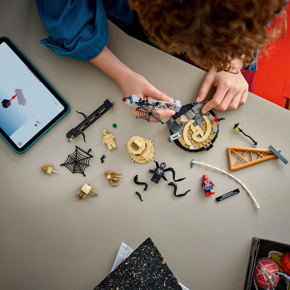 Osoba składająca zestaw Lego na stole. Rozrzucone elementy Lego, w tym figurki Spider-Mana, pajęczyny i konstrukcje.