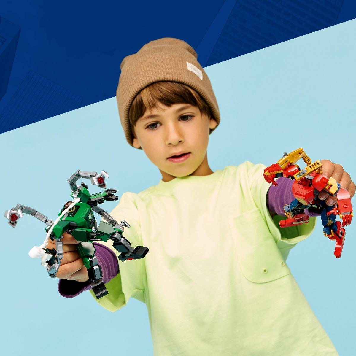 Chłopiec w czapce trzyma dwie zabawki robota, jedną zieloną z ramionami, drugą czerwoną i żółtą. Tło jest niebieskie.