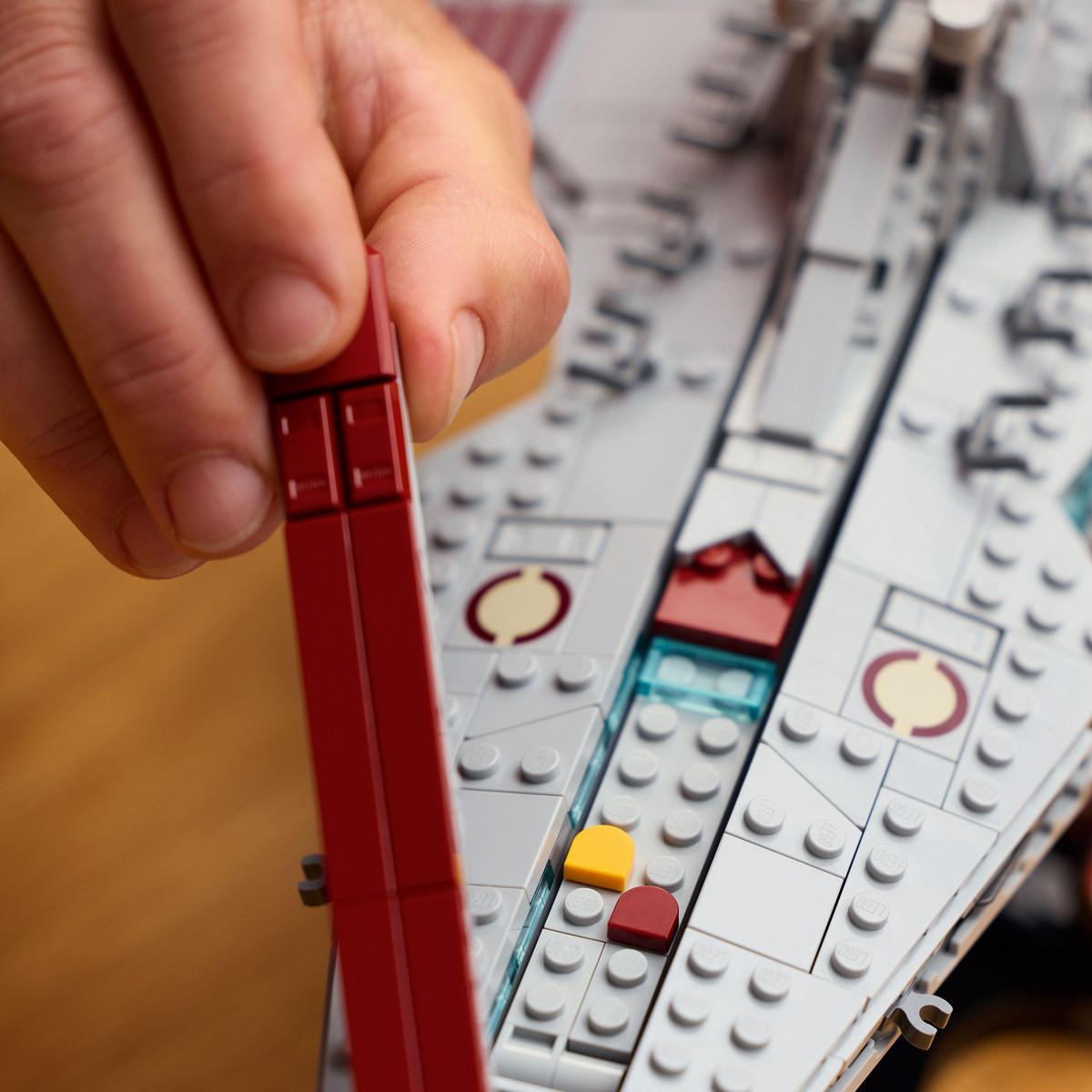 Ręka umieszcza czerwony klocek Lego na szarym modelu Lego. Model ma żółte i czerwone elementy. Tłem jest statek kosmiczny Lego.