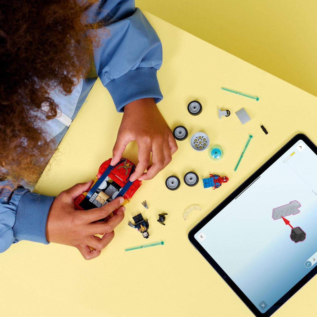 Dziecko składa czerwony samochód Lego z niebieskimi detalami, otoczone częściami, tabletem z instrukcjami i figurkami na żółtej powierzchni.