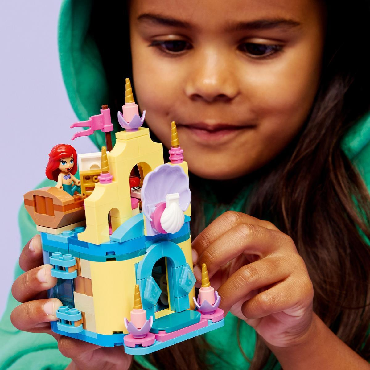 Una niña sostiene un juguete de castillo de Lego con una figura de Ariel. El castillo es amarillo, azul y rosa. La niña lleva una sudadera con capucha verde.